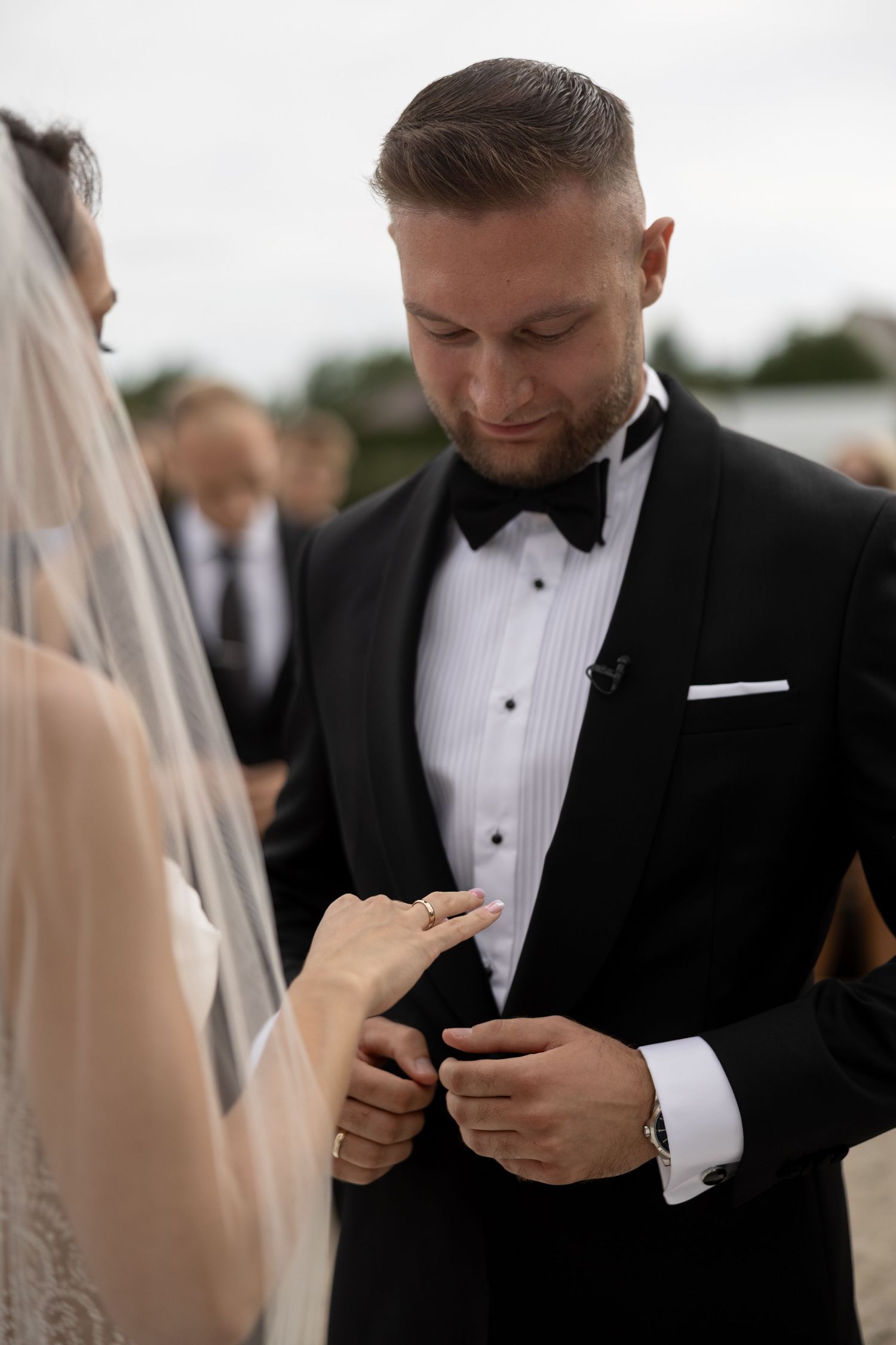 Groom in black tuxedo placing wedding ring on bride's finger during outdoor ceremony.