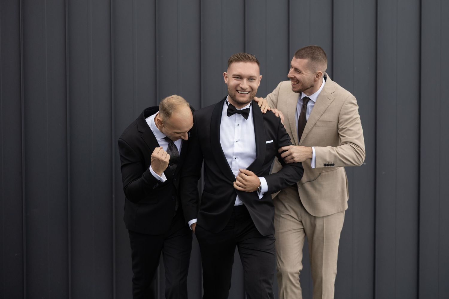 Three men in formal suits laughing together against a dark gray wall, groomsmen celebrating at a wedding.