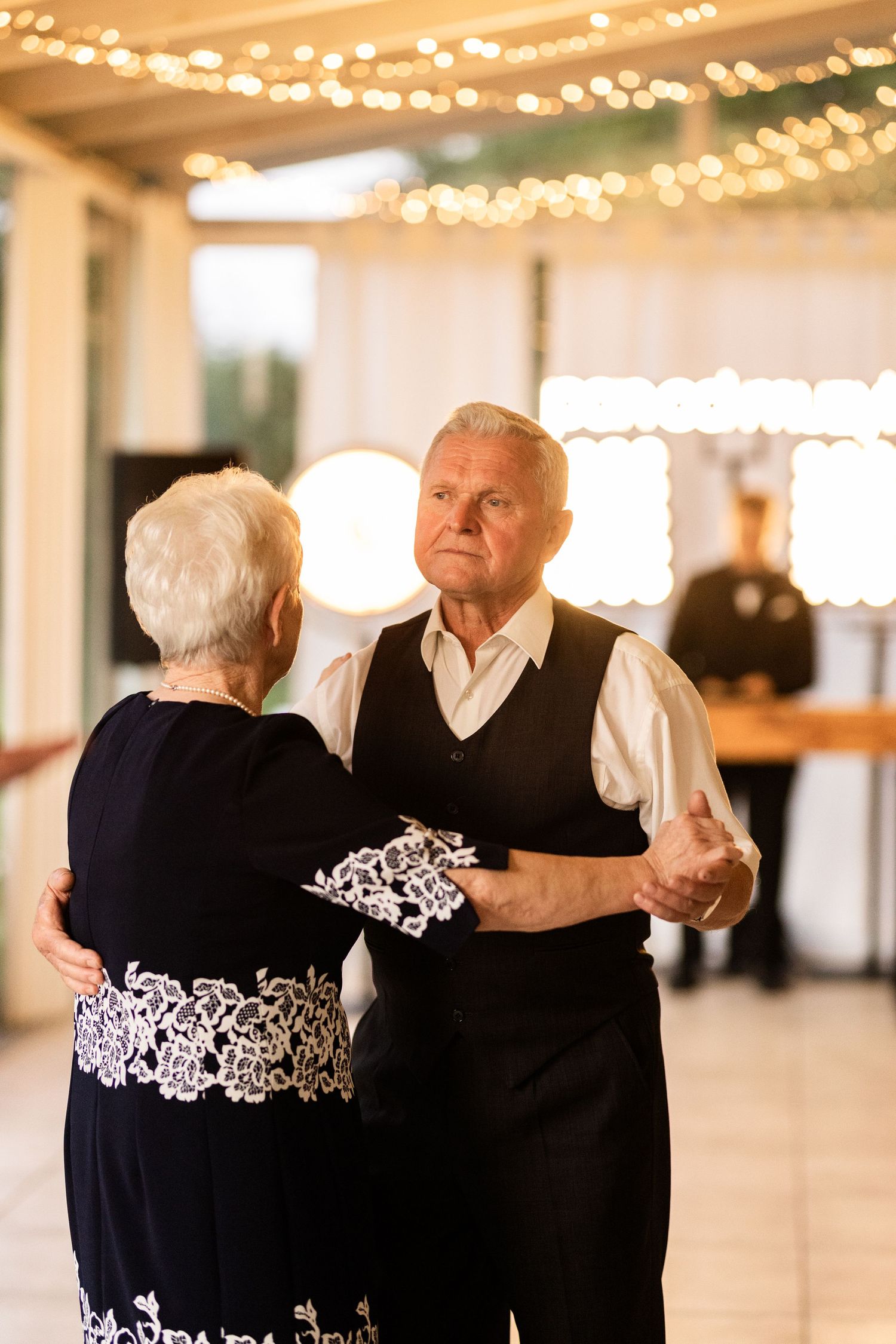 Elderly couple dancing together in a warmly lit ballroom with string lights overhead.