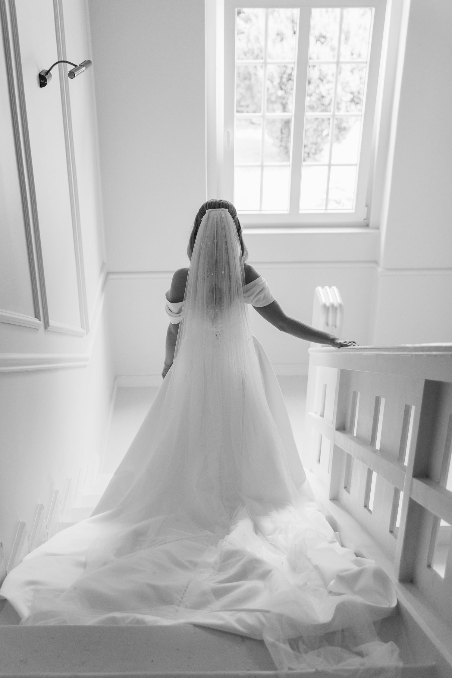 Black and white photo of a bride in a flowing wedding gown with long train standing by a bright window.