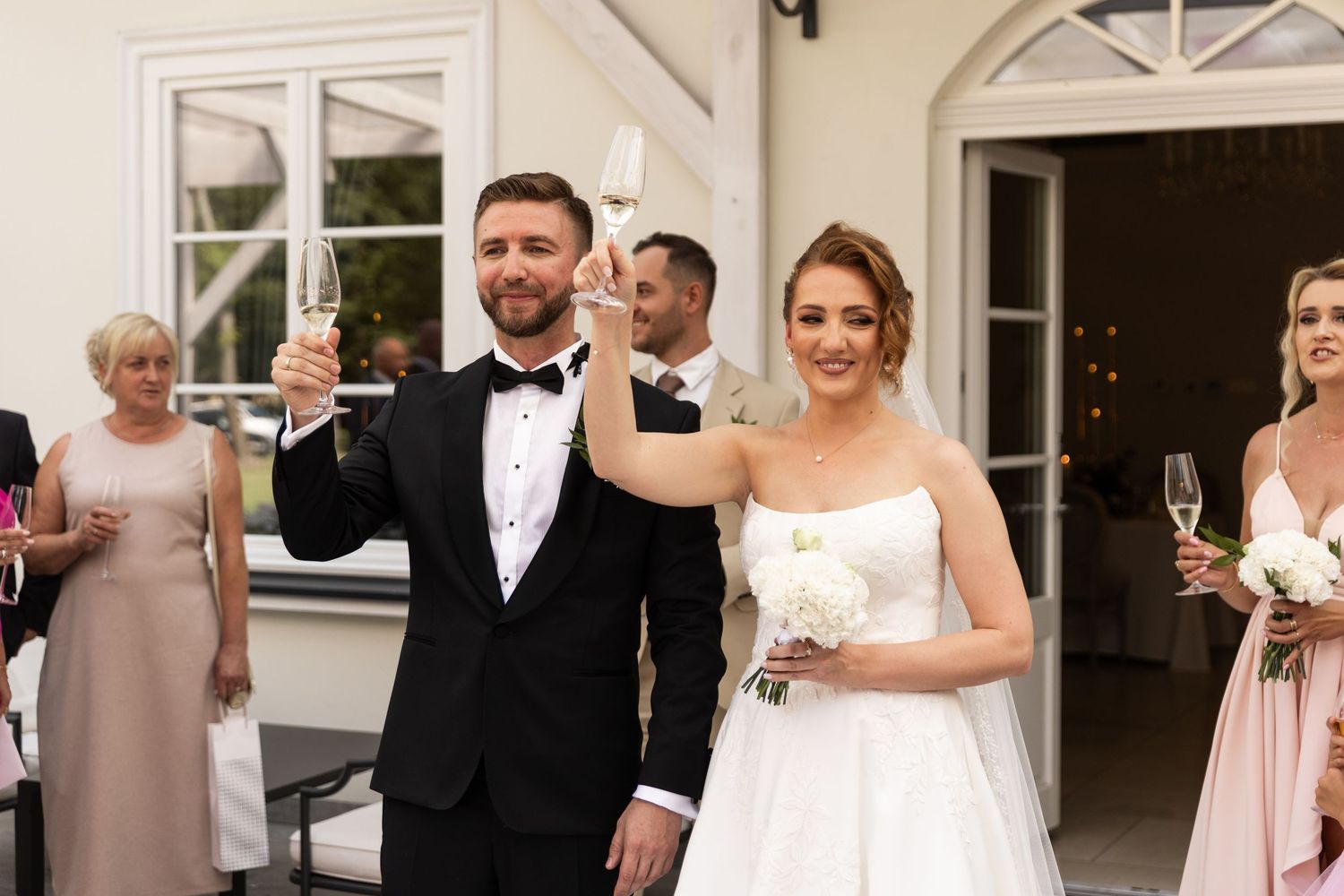 A wedding party poses in formal attire outside a white building with arched doorways and windows.