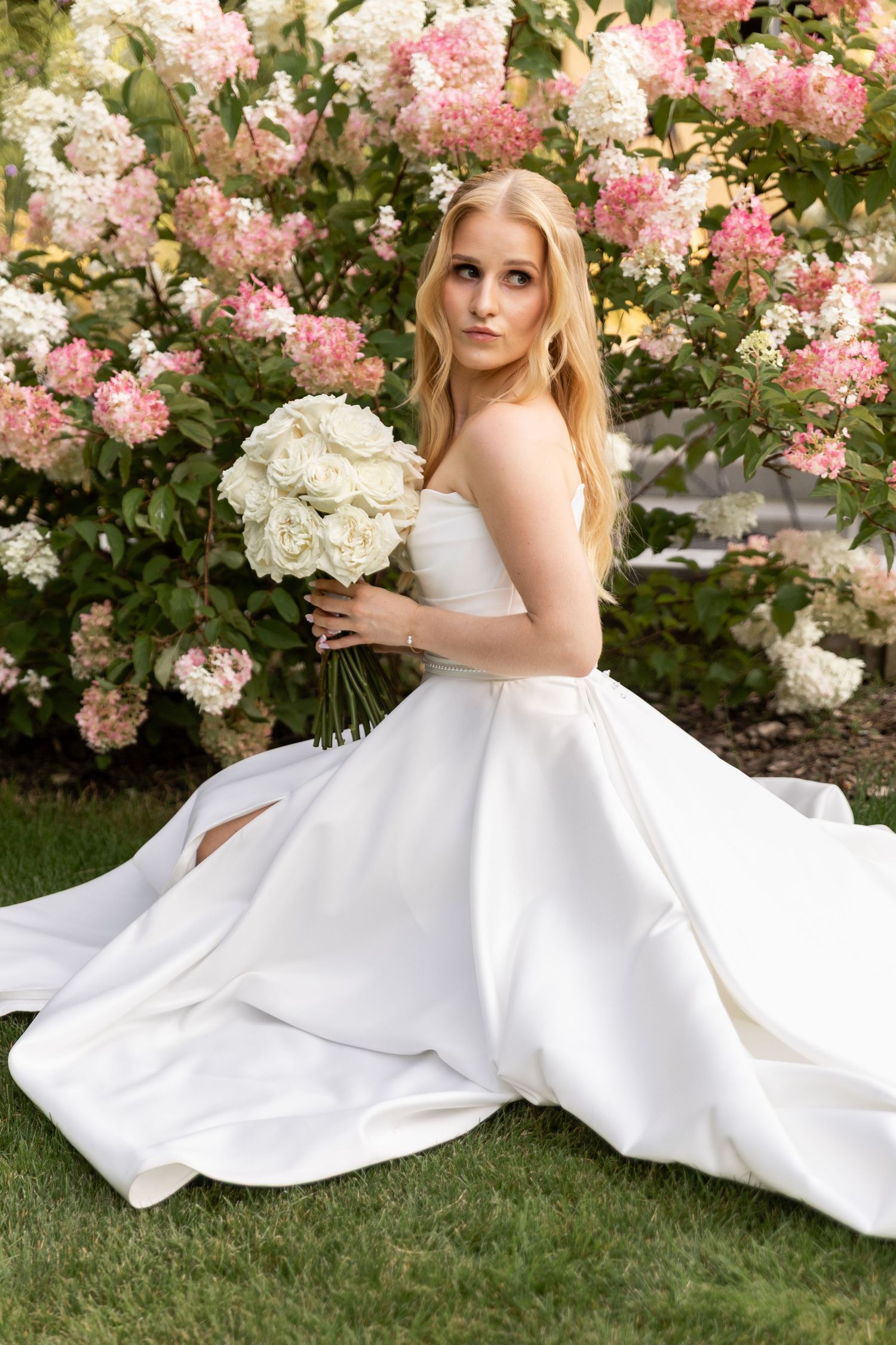 A bride in a flowing white gown poses gracefully on grass near blooming pink and white hydrangea bushes.