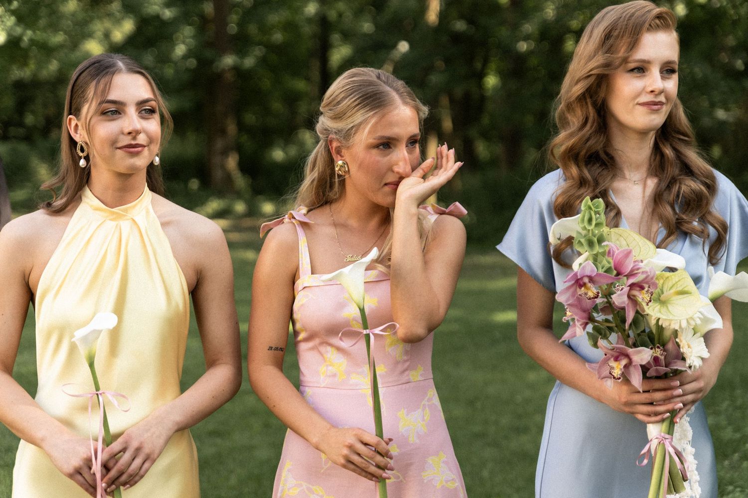 Three bridesmaids in pastel dresses stand outdoors holding single-stem flowers at a summer wedding ceremony.