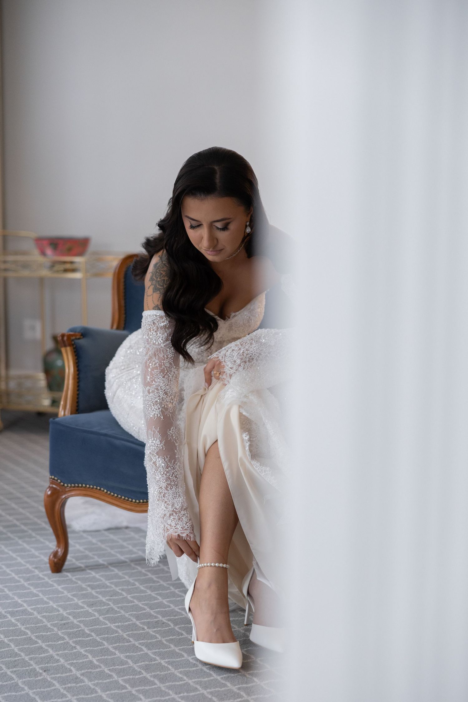 Bride in white lace robe fastening pearl ankle strap heels while seated on blue velvet chair before wedding ceremony.