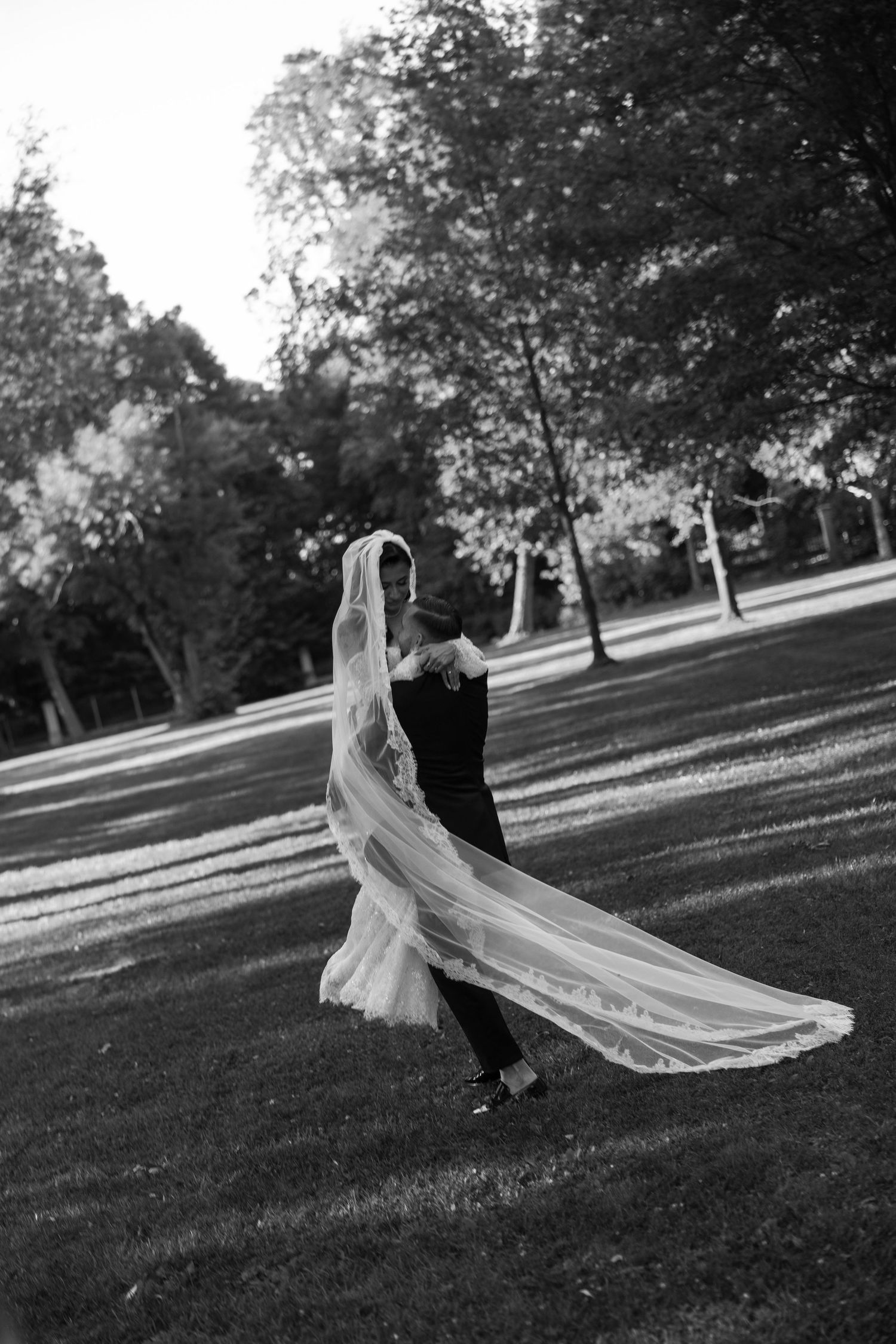 Black and white photo of bride and groom dancing in a park, her long veil flowing in the wind.
