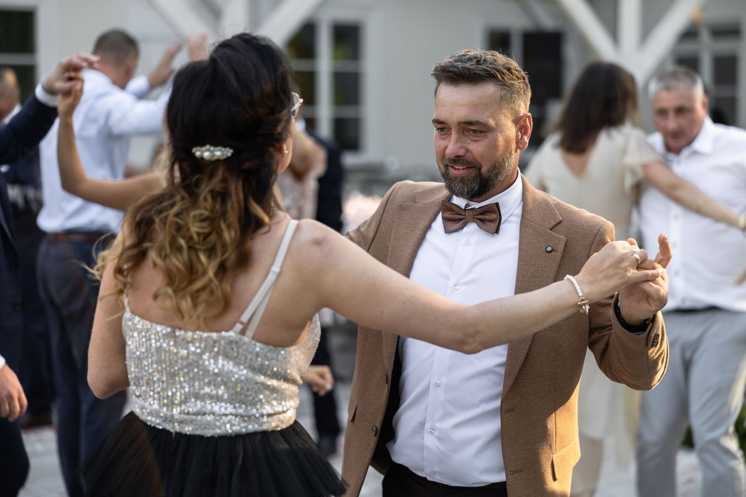 Elegant couple dancing at a formal outdoor event, man in tan blazer and bow tie, woman in sequined dress.