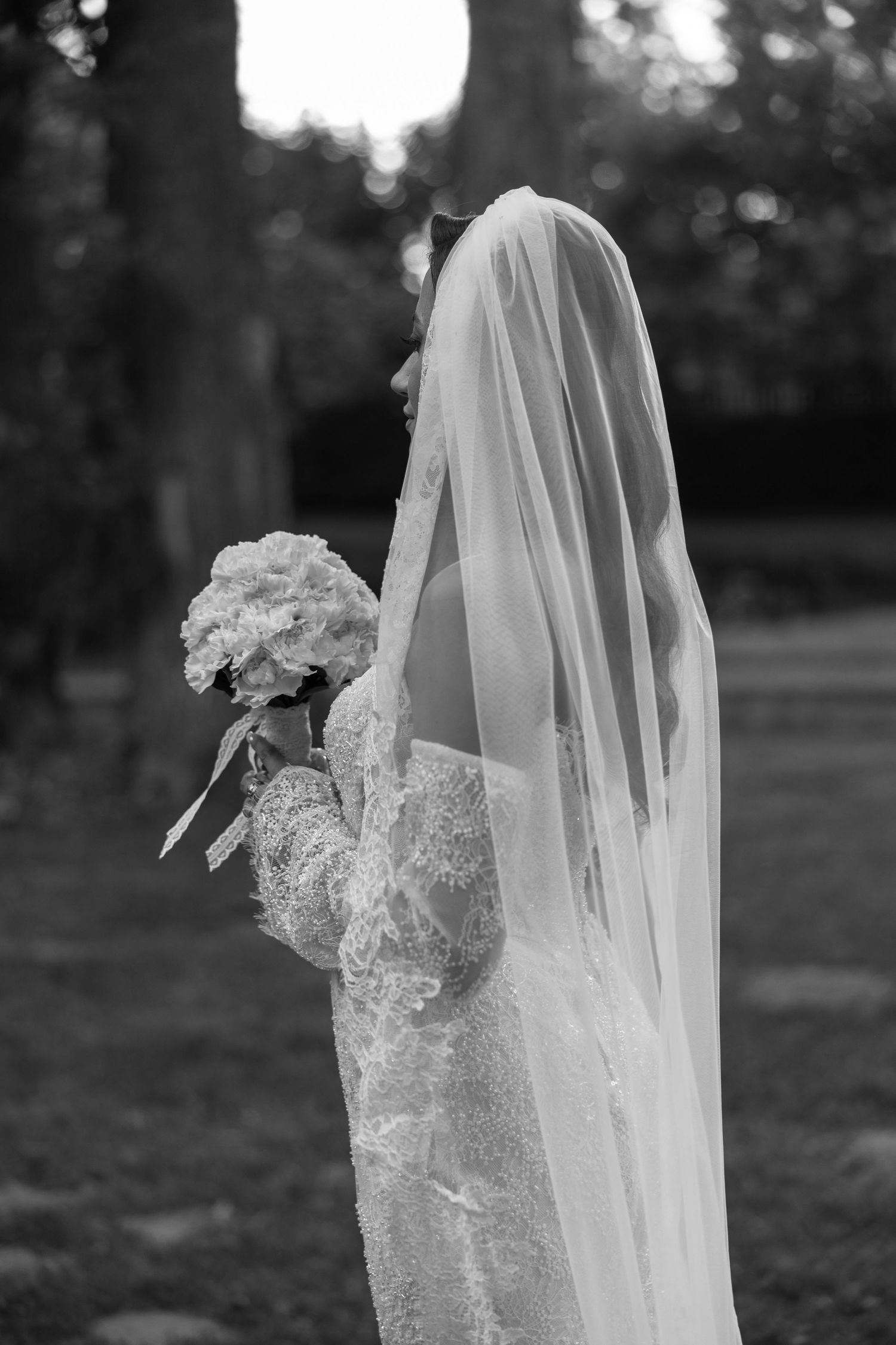Black and white photo of a bride in lace gown and veil holding a white bouquet outdoors.