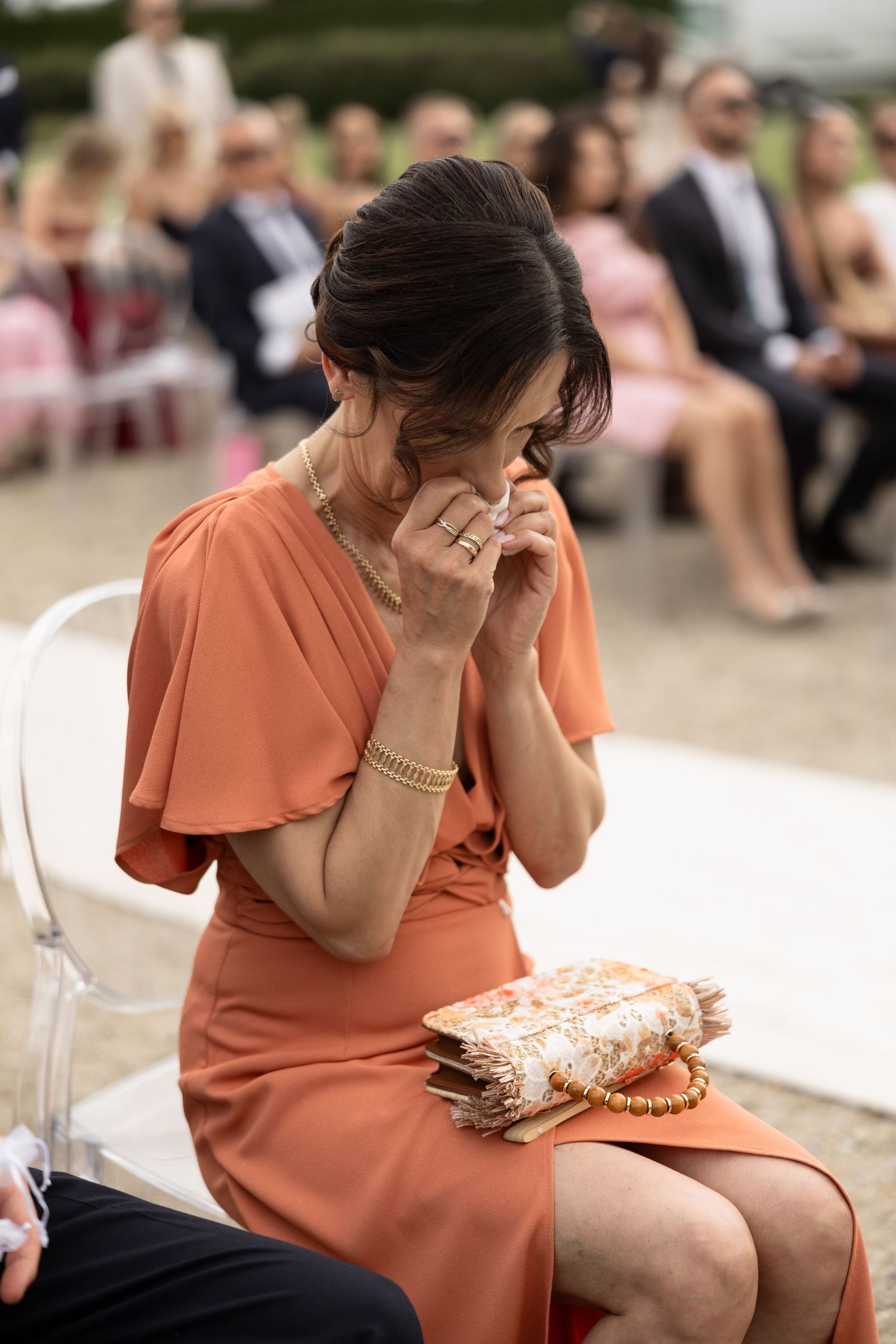 Emotional wedding guest in orange dress wiping tears, holding floral clutch at outdoor ceremony.