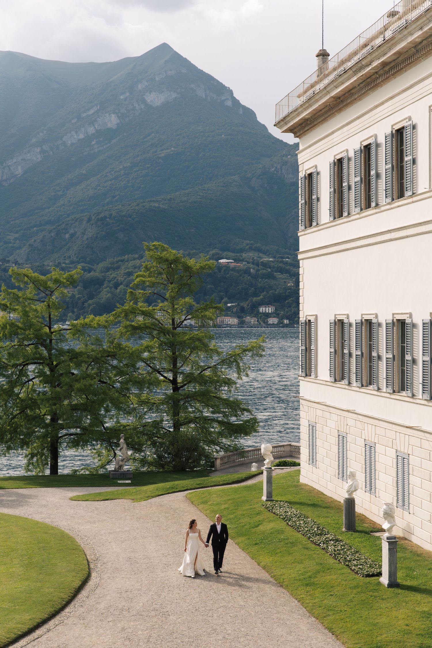 Bride and groom walking hand in hand along a path by a grand villa on Lake Como, Italy, with mountains in the background.