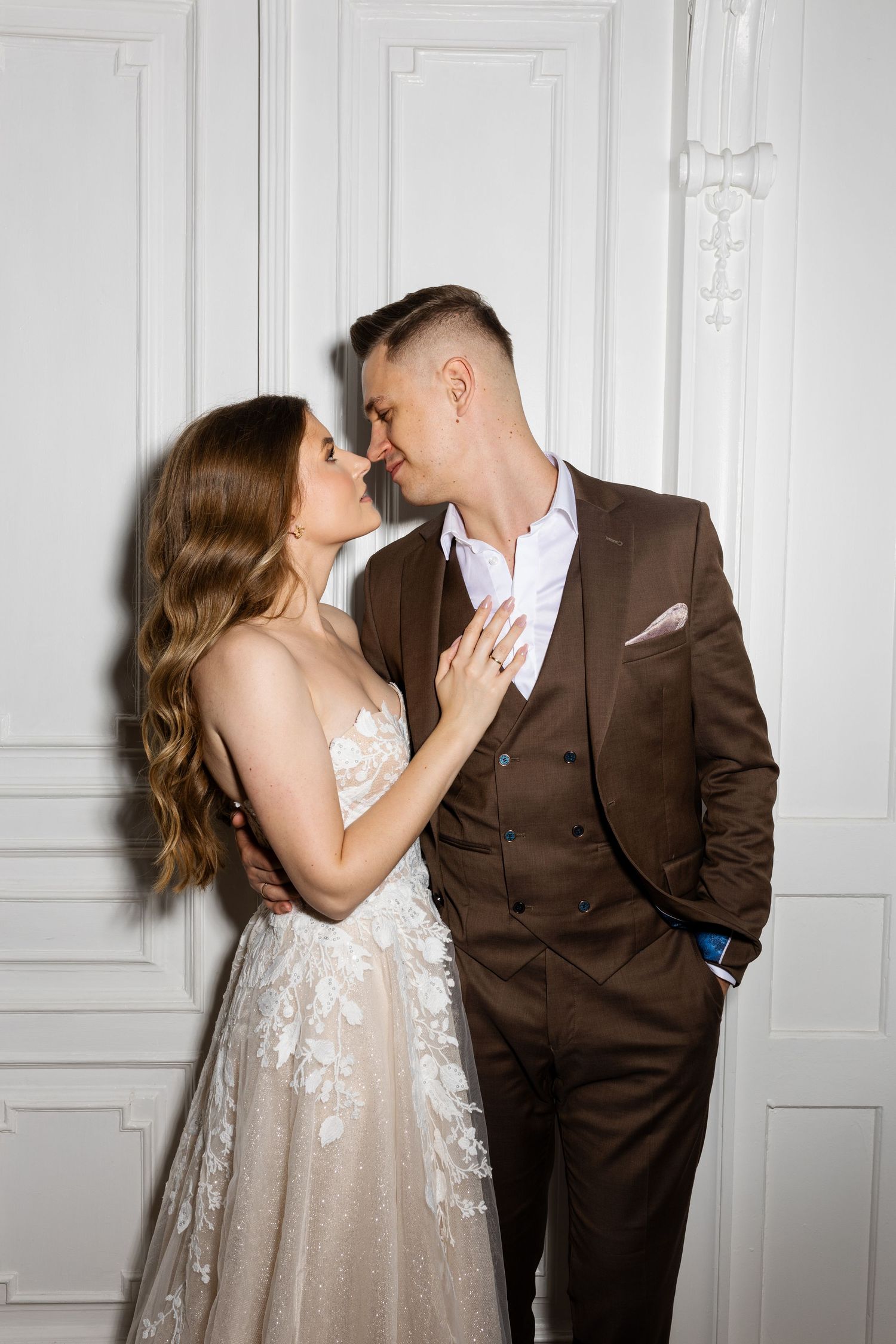 Intimate wedding portrait with couple in brown suit and lace dress against white paneled wall.