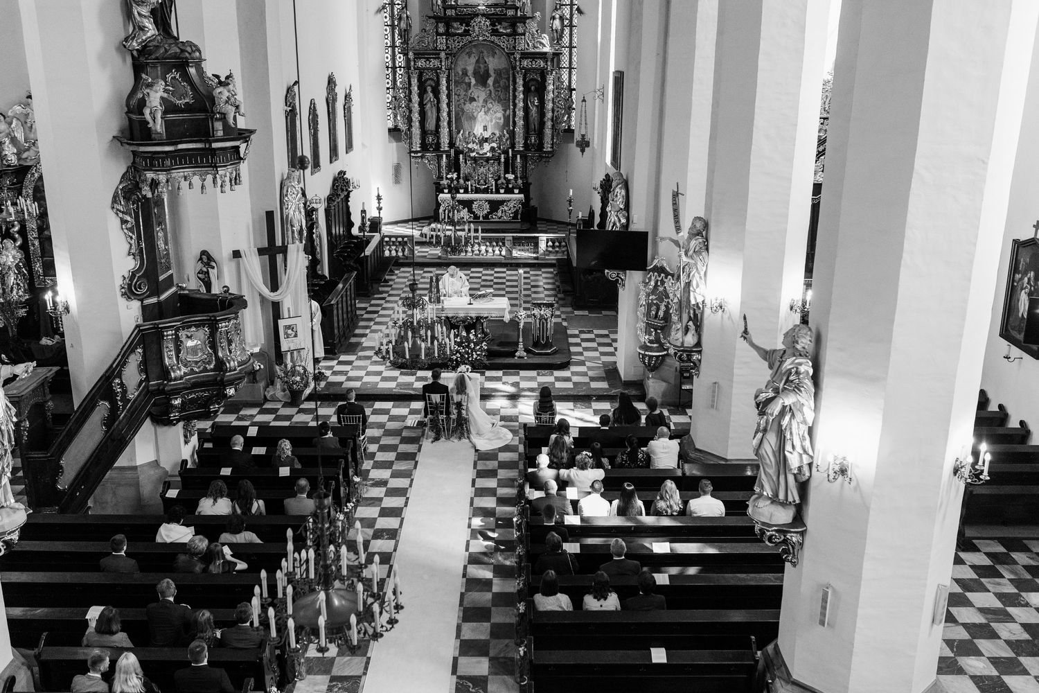 High-angle black and white view of church interior with checkered floor and people seated in wooden pews during service.