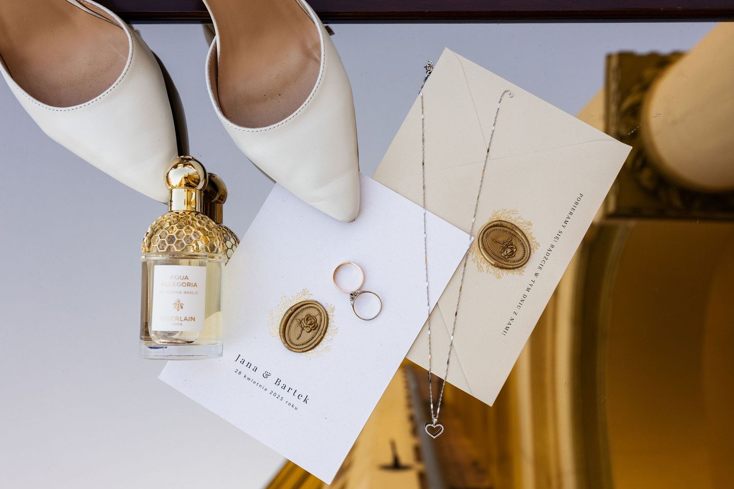 Wedding accessories including white stiletto heels, perfume bottle, wedding rings, and invitation cards with wax seals on white surface.
