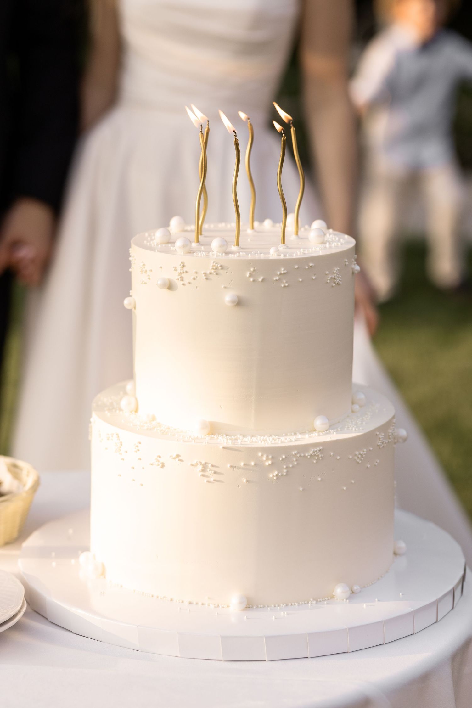 White wedding cake with tall gold candles displayed in multiple angles on a white cake stand at an outdoor reception.