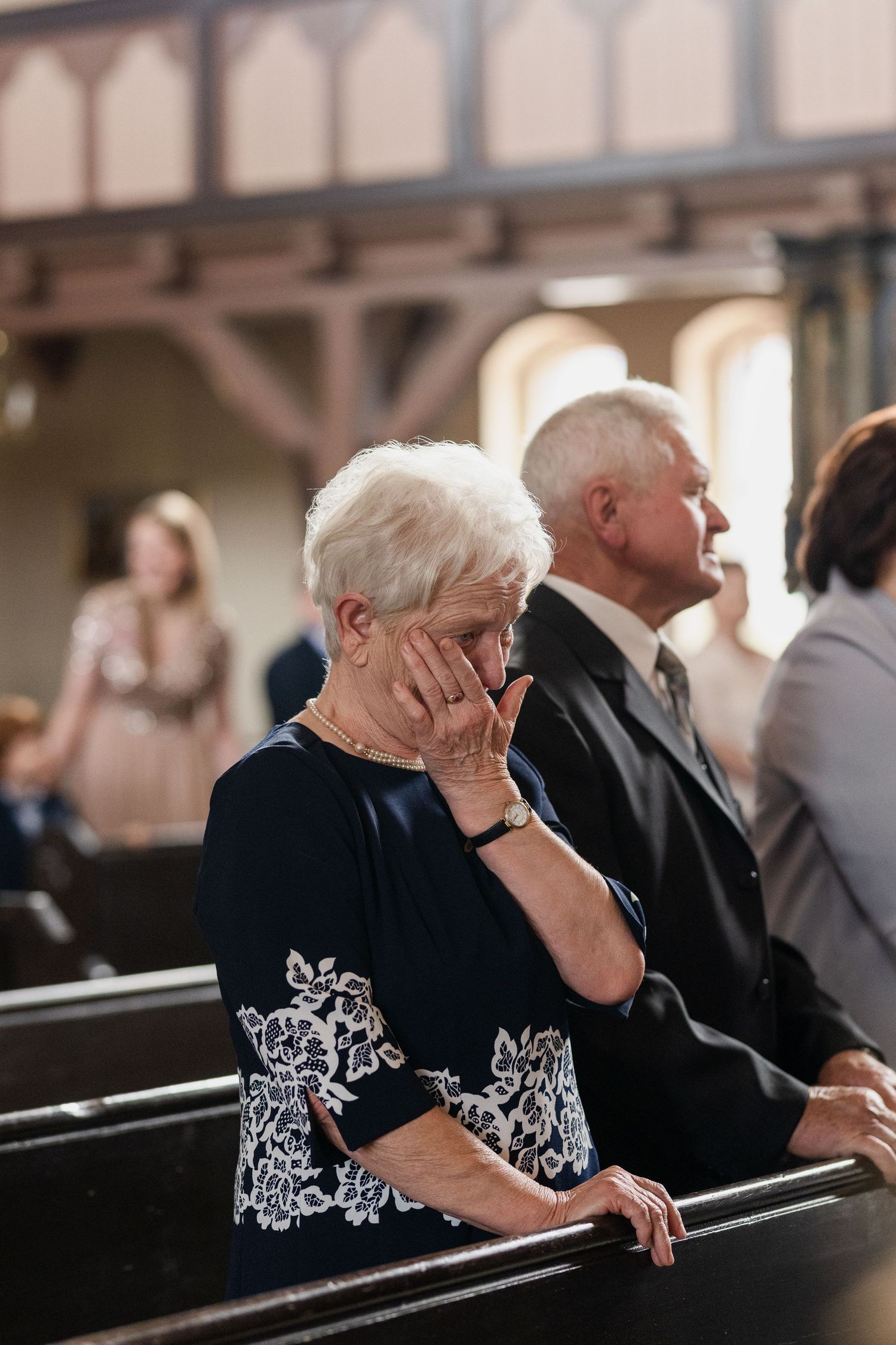 A person in a black floral top sits in a church pew with their hand resting against their face during a service.