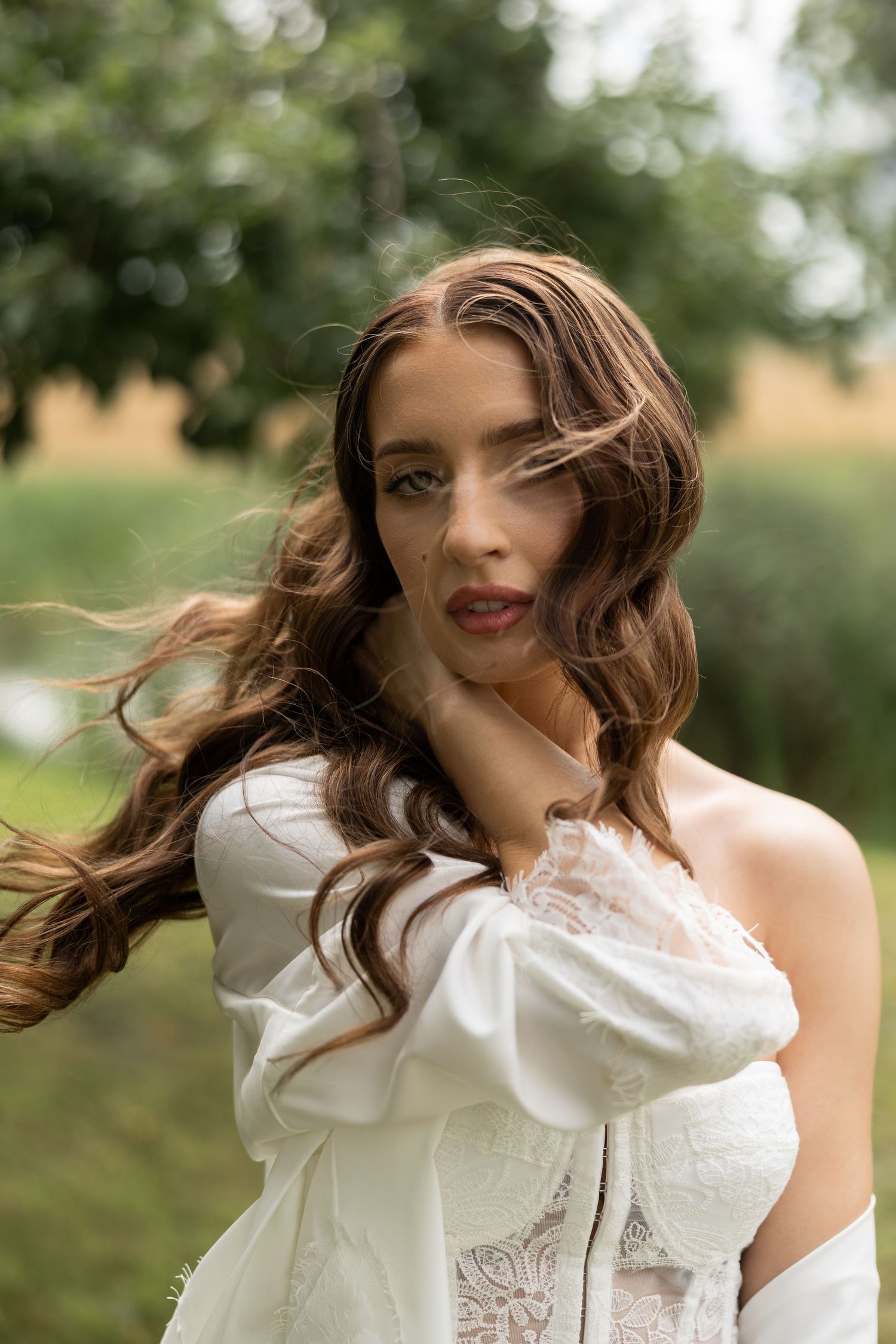 Young woman with long wavy brown hair in white lace dress posing outdoors with green trees in background.