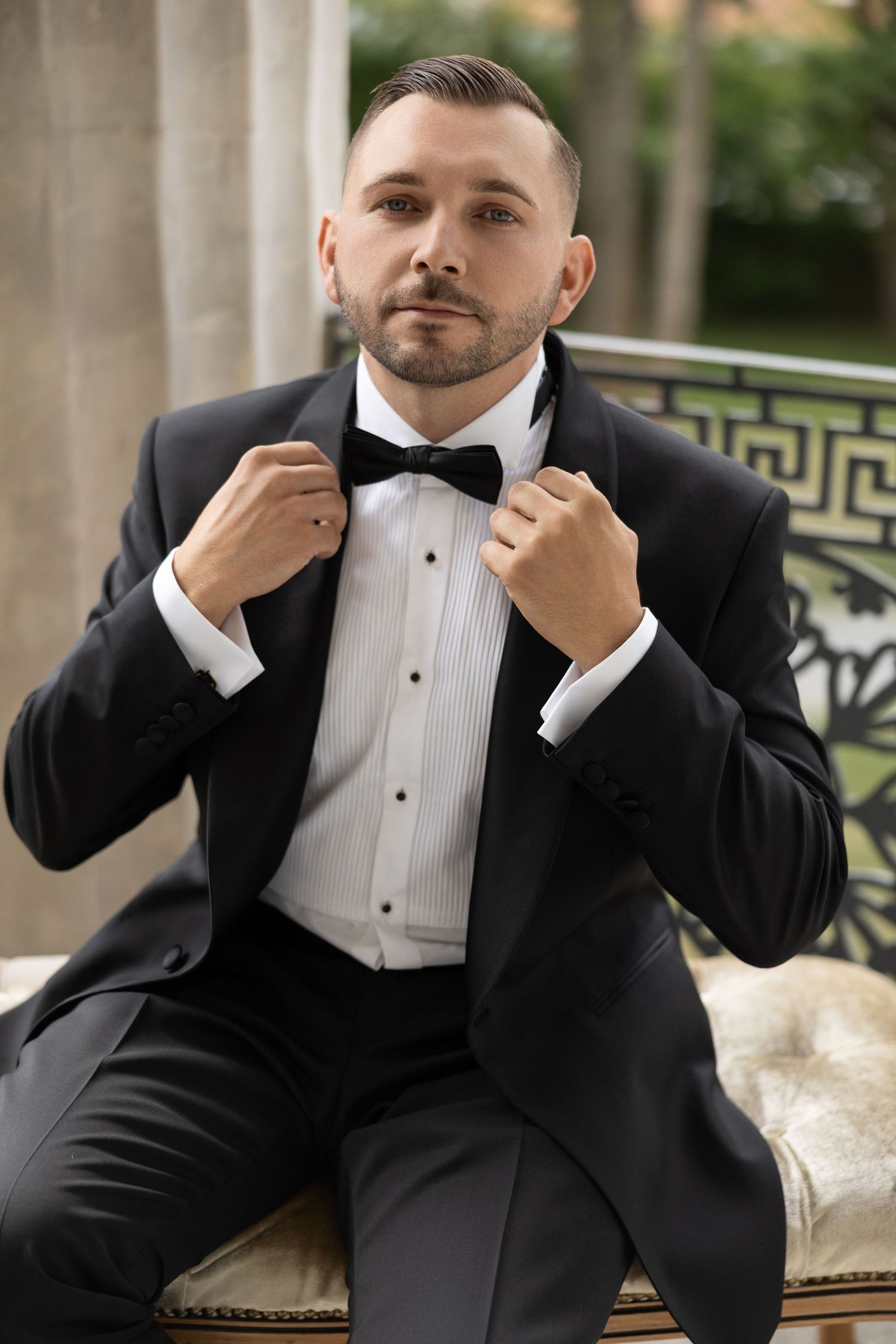 A well-dressed man in a black tuxedo and bow tie sits confidently, adjusting his collar in an elegant outdoor setting.