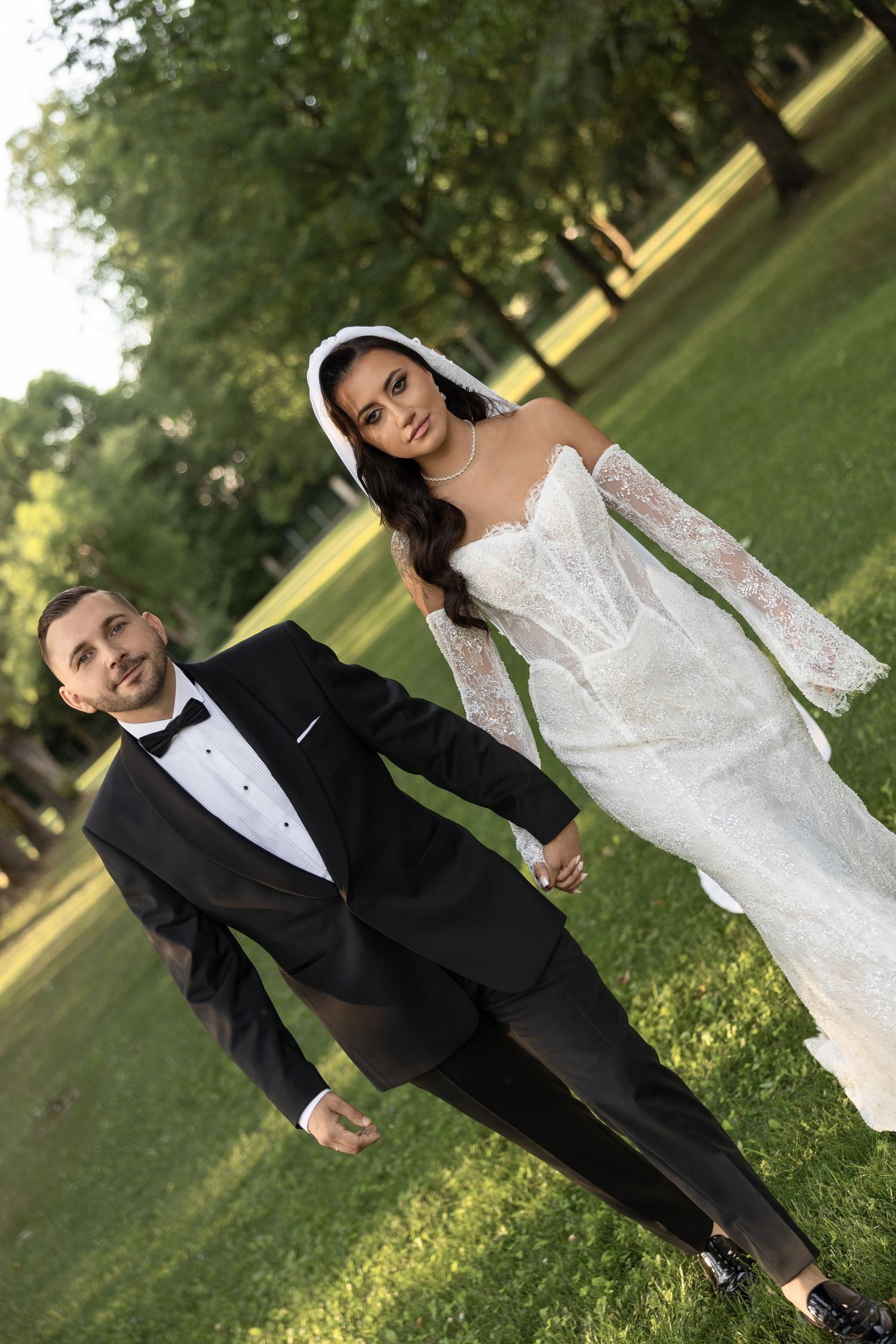 A bride in a white lace gown and veil holds hands with a groom in a black tuxedo in a lush green park.
