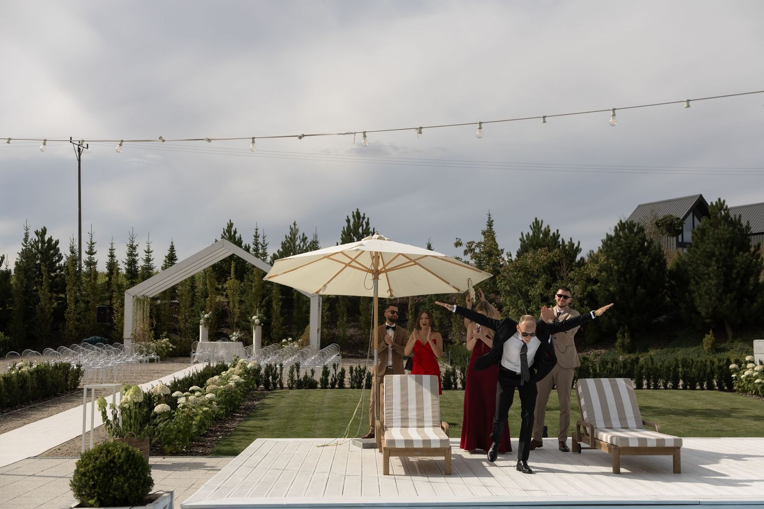 Elegant outdoor wedding venue with string lights, white tent, umbrella, lounge chairs, and guests posing near lush greenery.