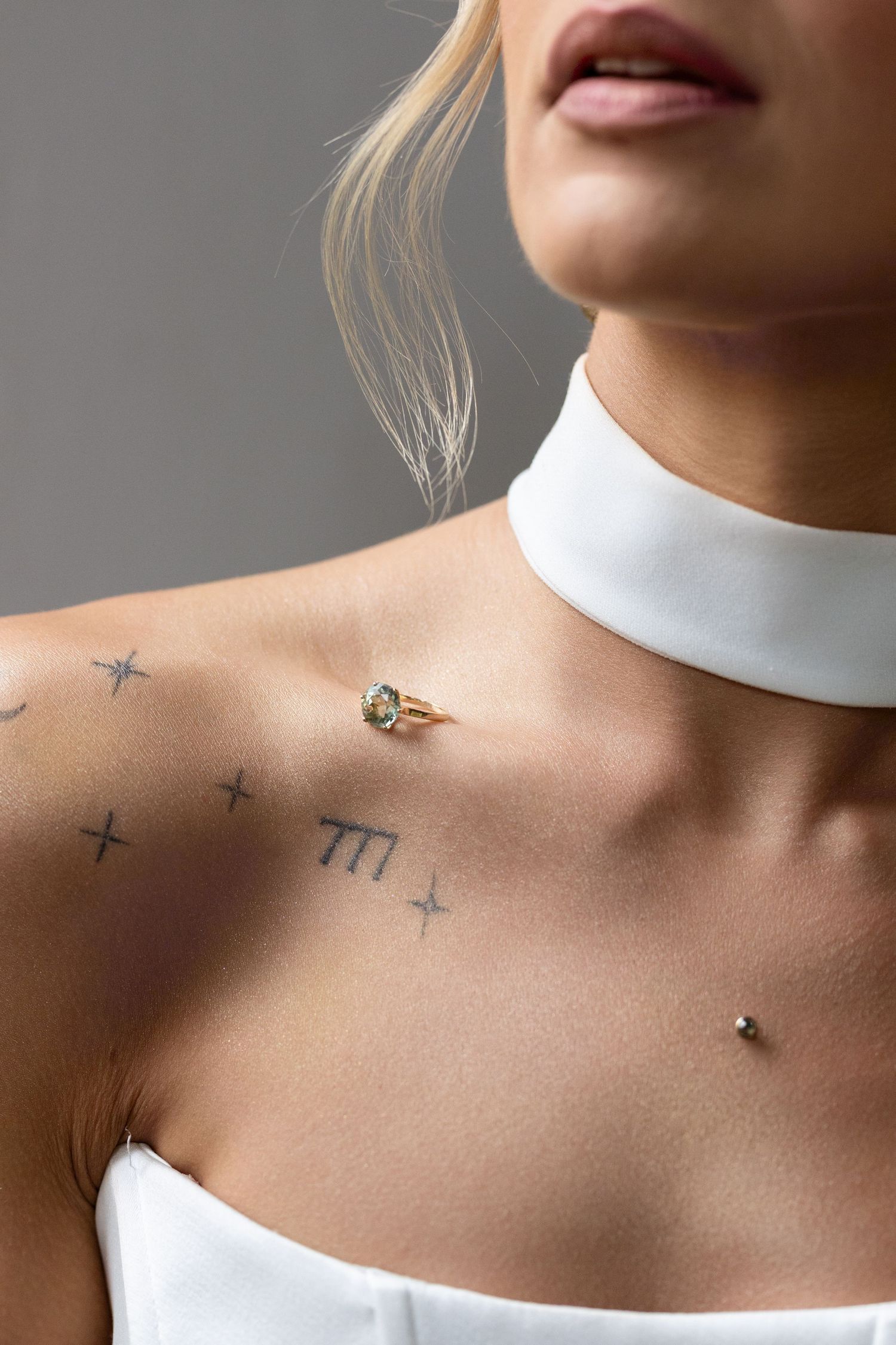 Woman with star tattoos on chest wearing white choker and gold ring, blonde hair, gray background.