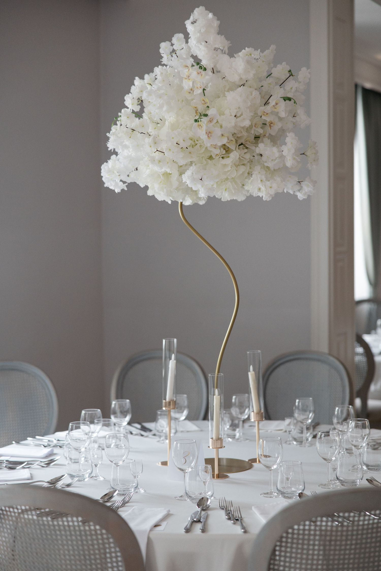 Elegant wedding table centerpiece with white floral arrangement on gold stand, surrounded by crystal glassware and candles.