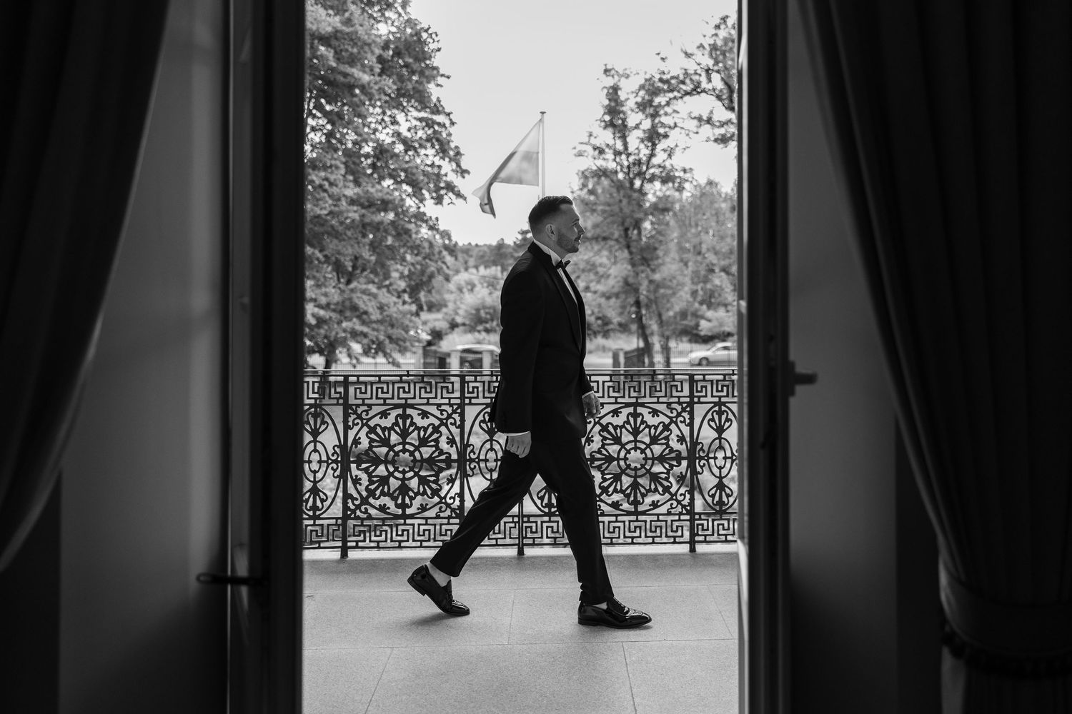 Black and white photo of a man in a suit walking on an ornate balcony with a flag visible in the background.