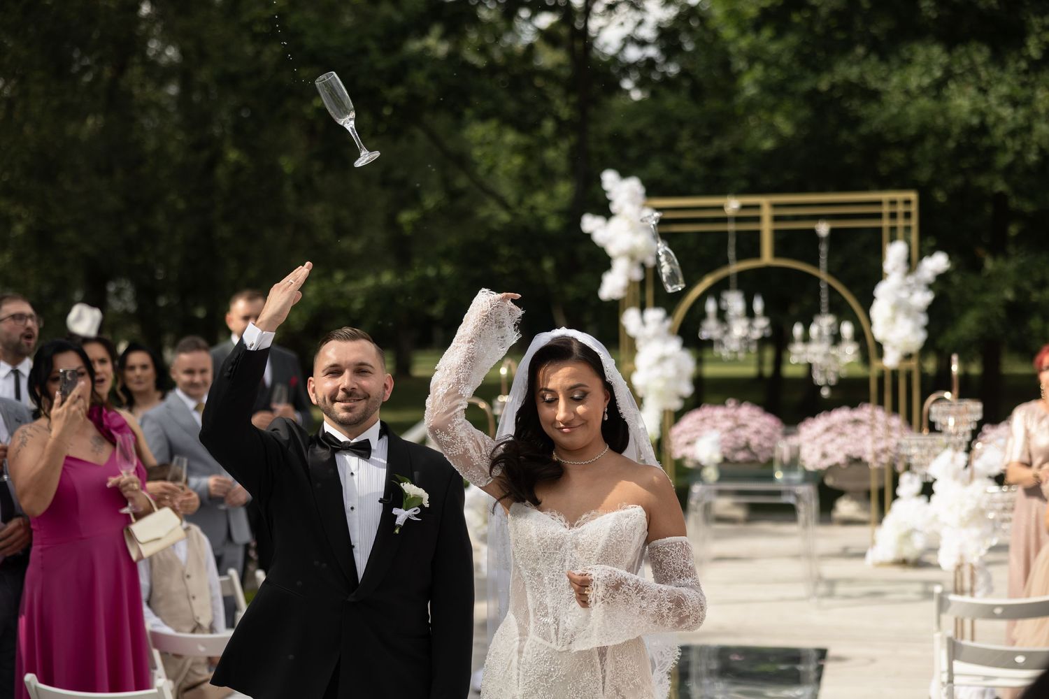 Joyful newlyweds celebrate outdoors raising champagne glasses after wedding ceremony with floral arch decor.