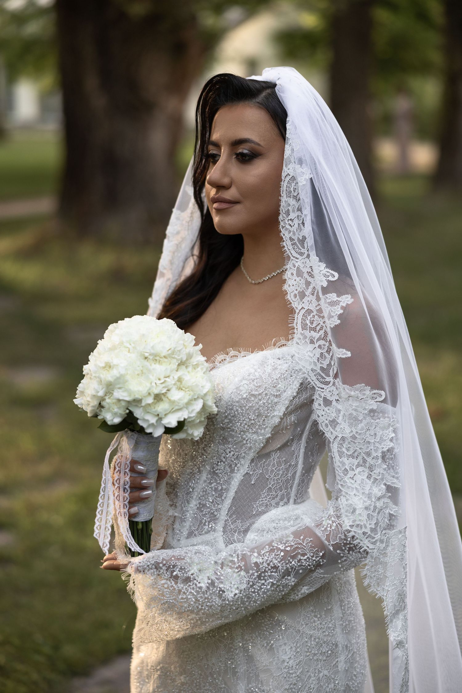 Elegant bride in lace wedding dress and veil holding white hydrangea bouquet outdoors.