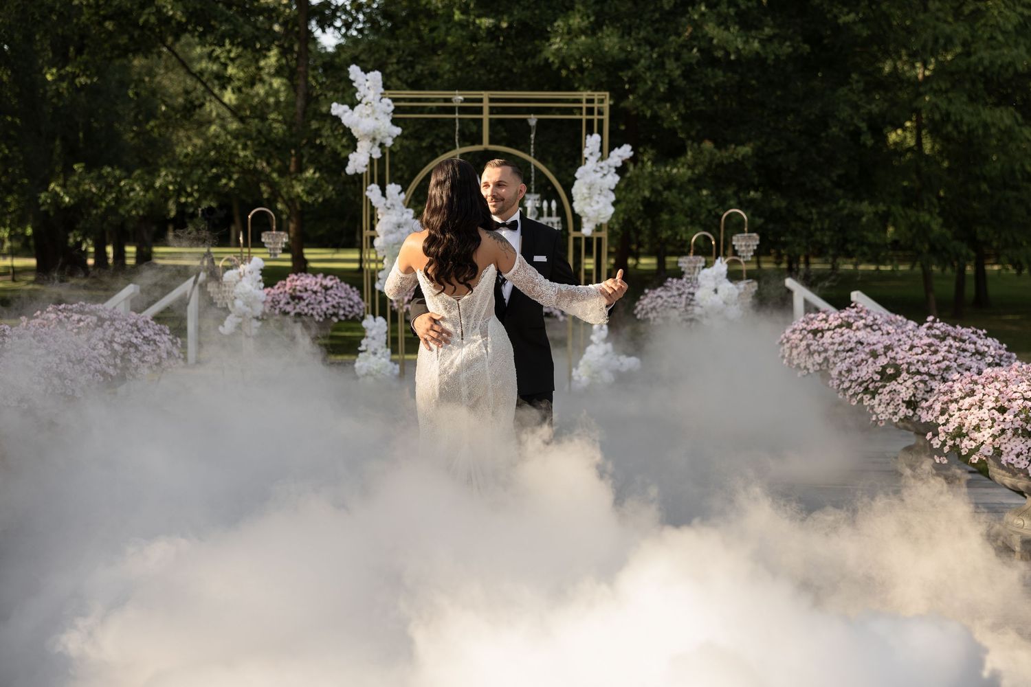 Bride and groom emerge through dramatic fog at outdoor wedding ceremony with floral arch and white balloon decor.