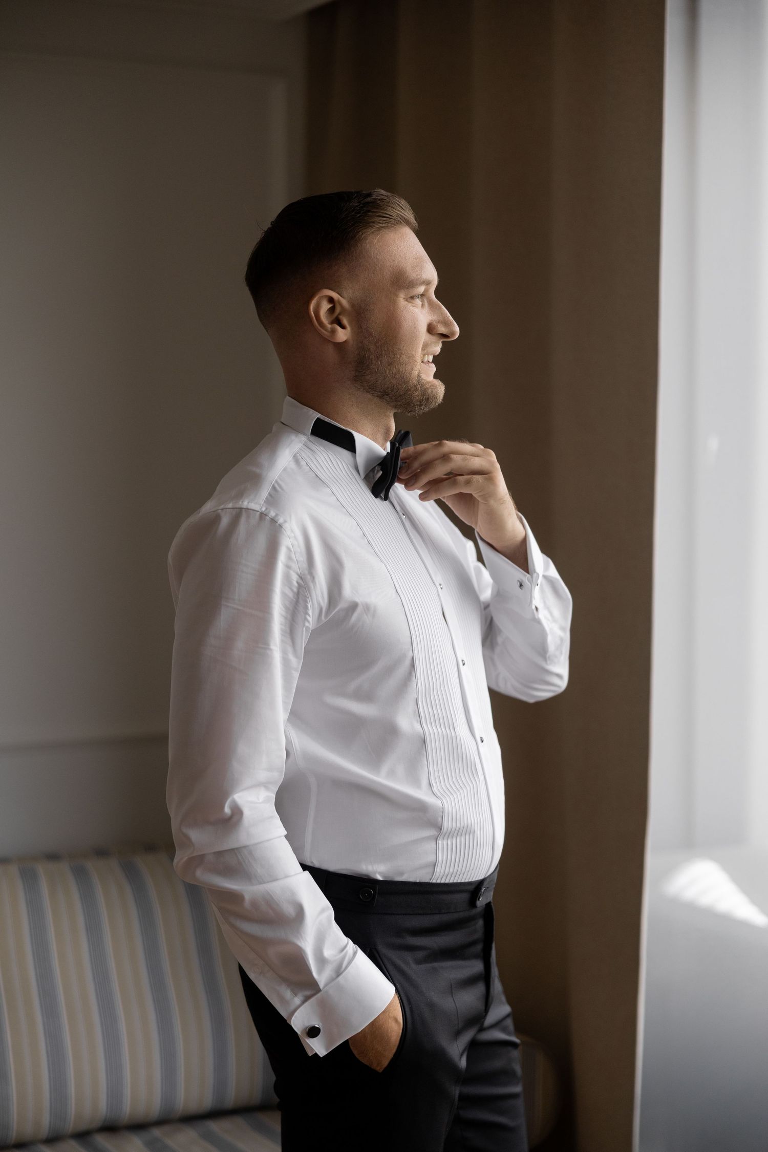 A well-dressed groom in a white tuxedo shirt adjusts his black bow tie while gazing out a window.