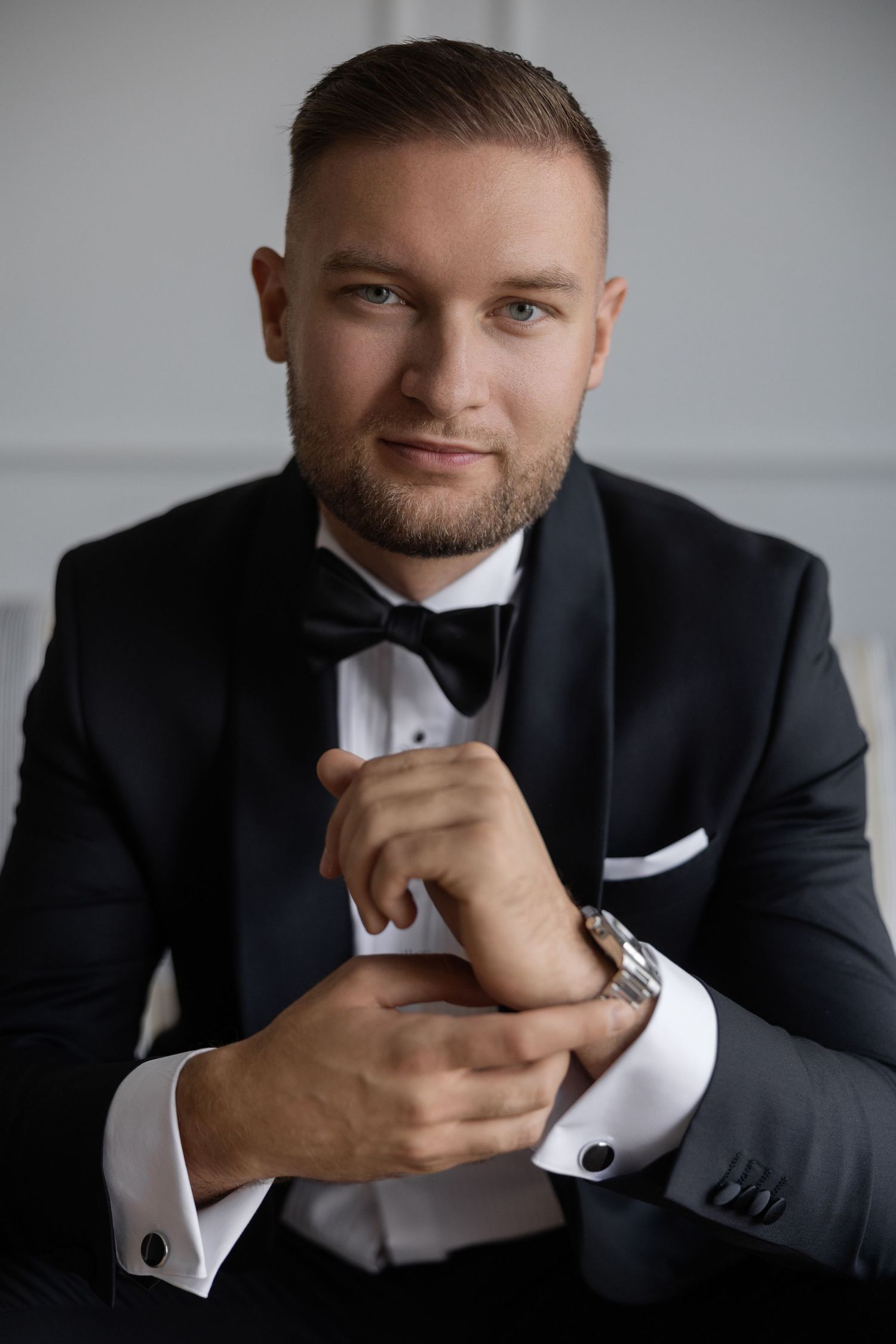 Elegant man in black tuxedo with bow tie adjusting cufflinks, smiling confidently in a formal portrait setting.