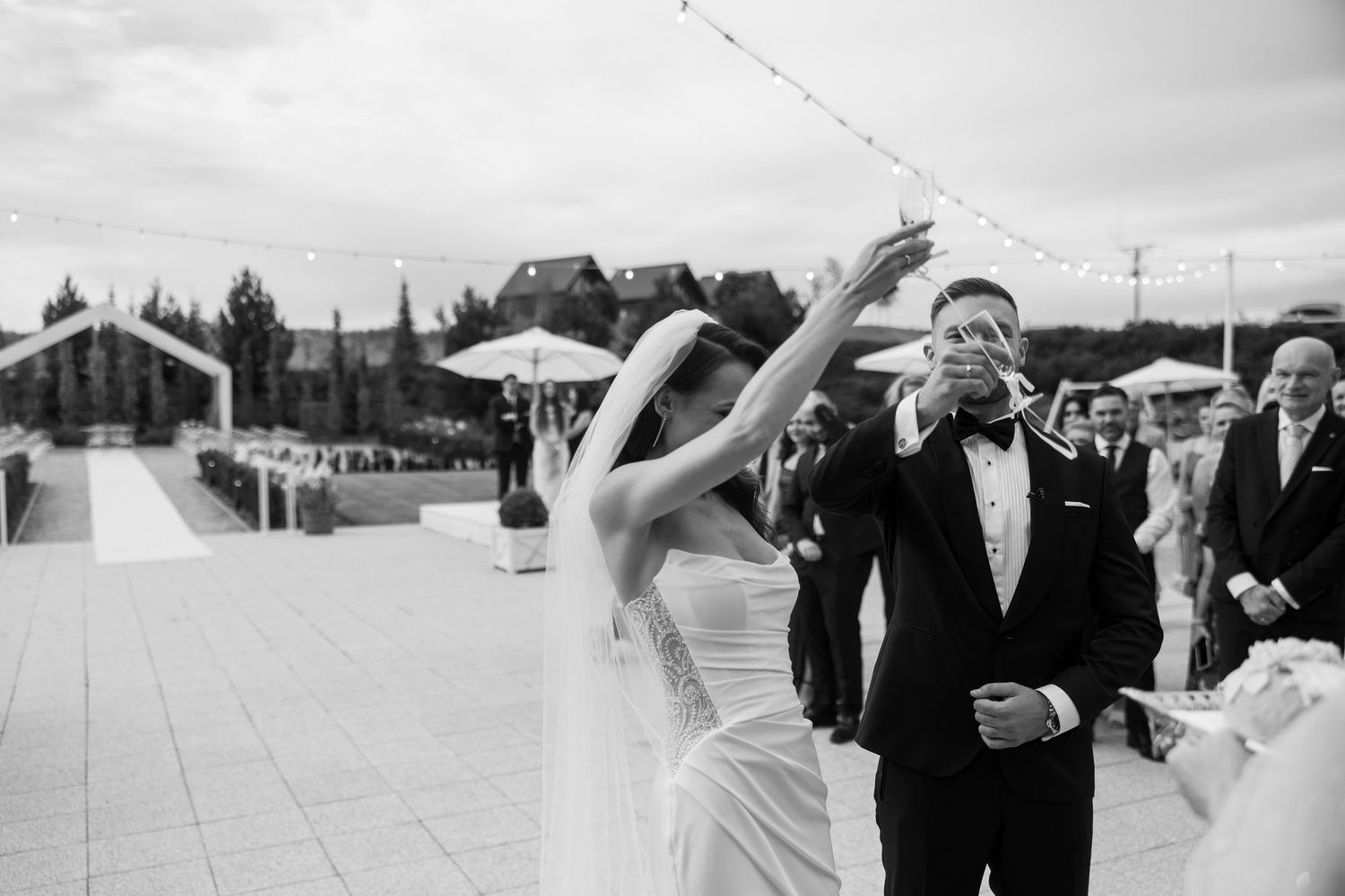Black and white photo of bride and groom celebrating outdoors at elegant wedding venue with string lights.