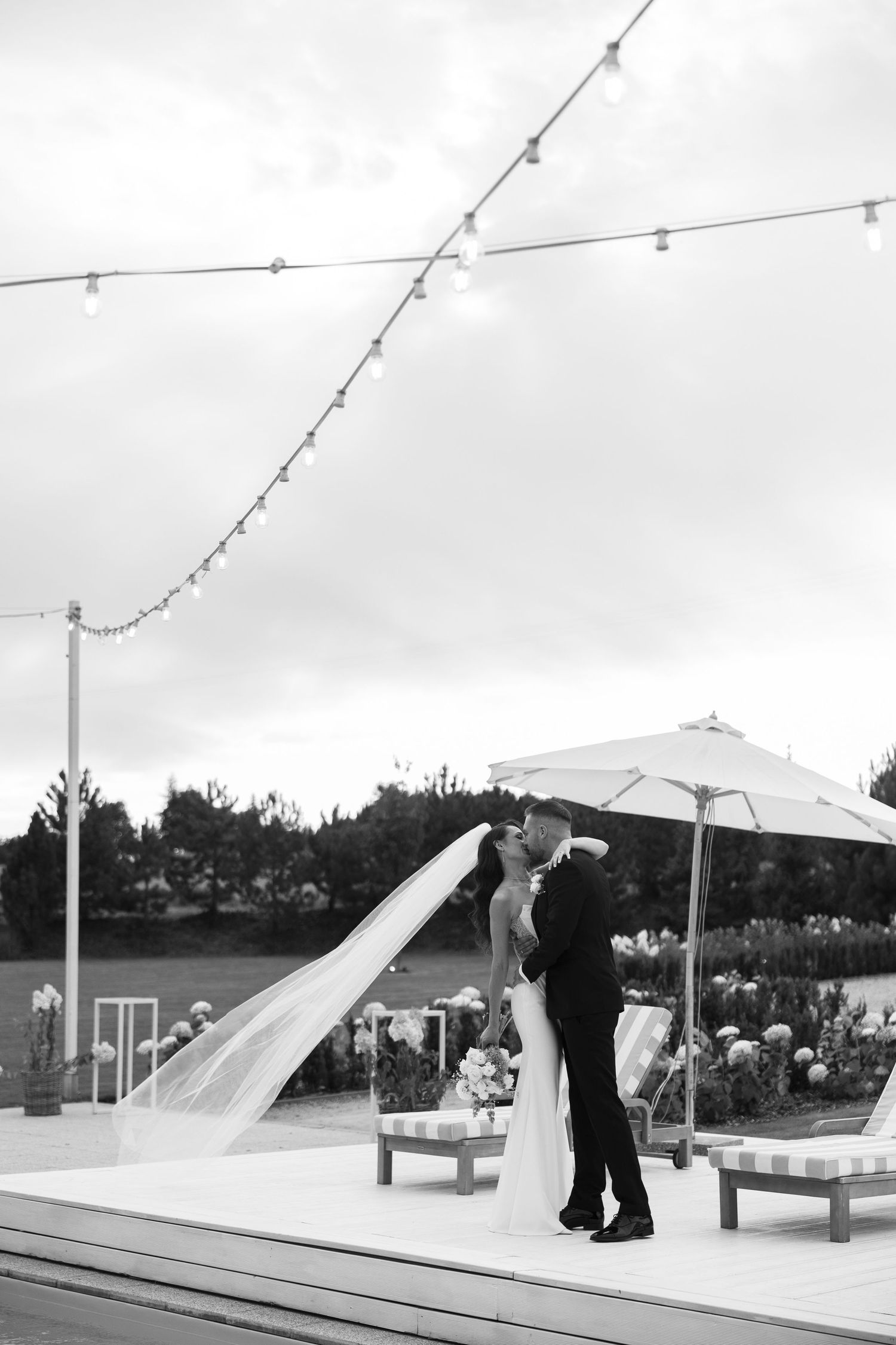 Black and white photo of a bride and groom sharing a romantic kiss outdoors under string lights at their wedding.