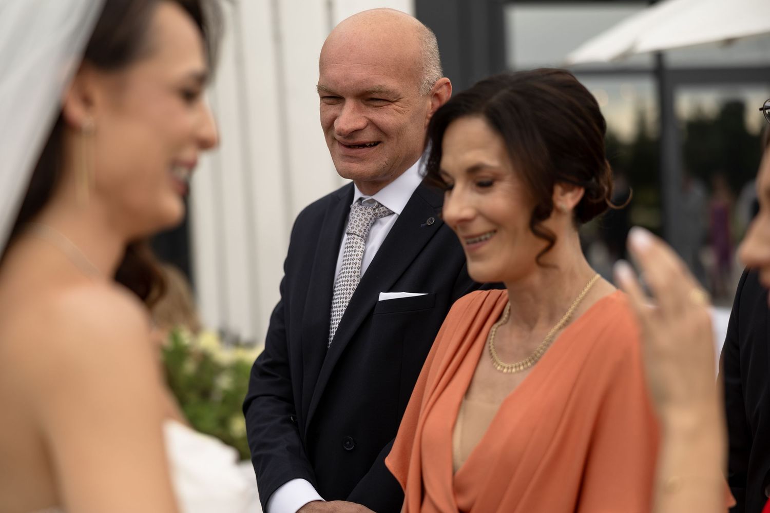 A bride in white dress smiles with elegantly dressed wedding guests at an outdoor ceremony.