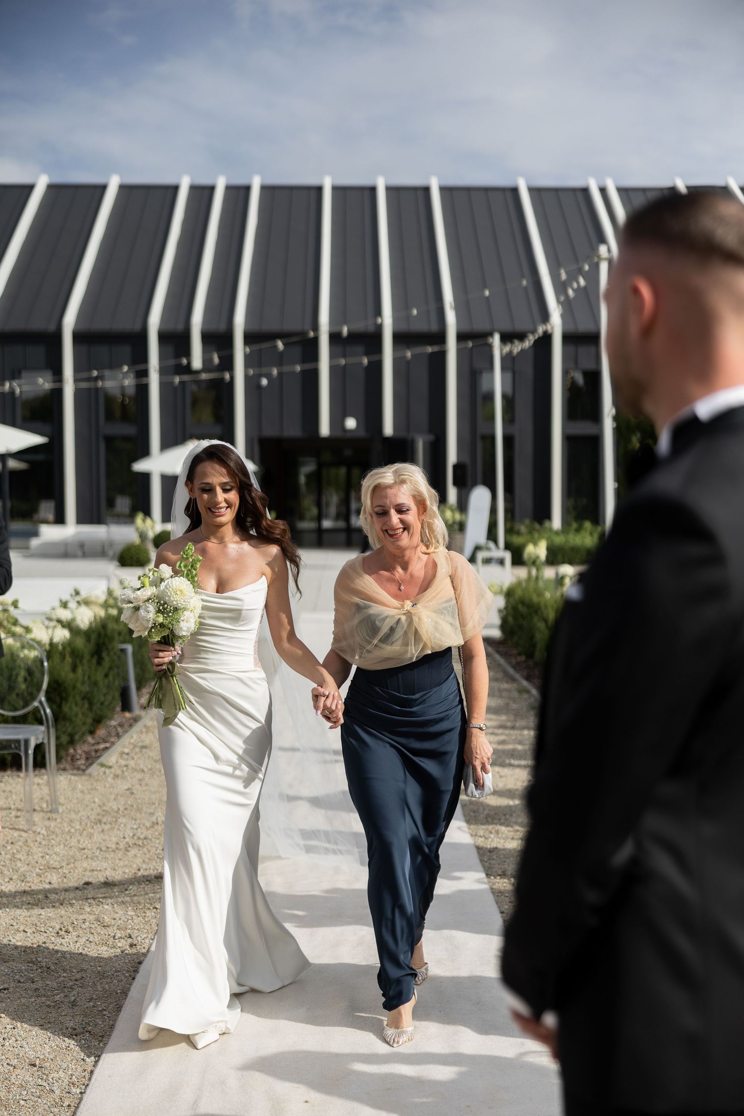 Bride in white gown walks down outdoor aisle with older woman, holding bouquet, as groom waits in tuxedo.