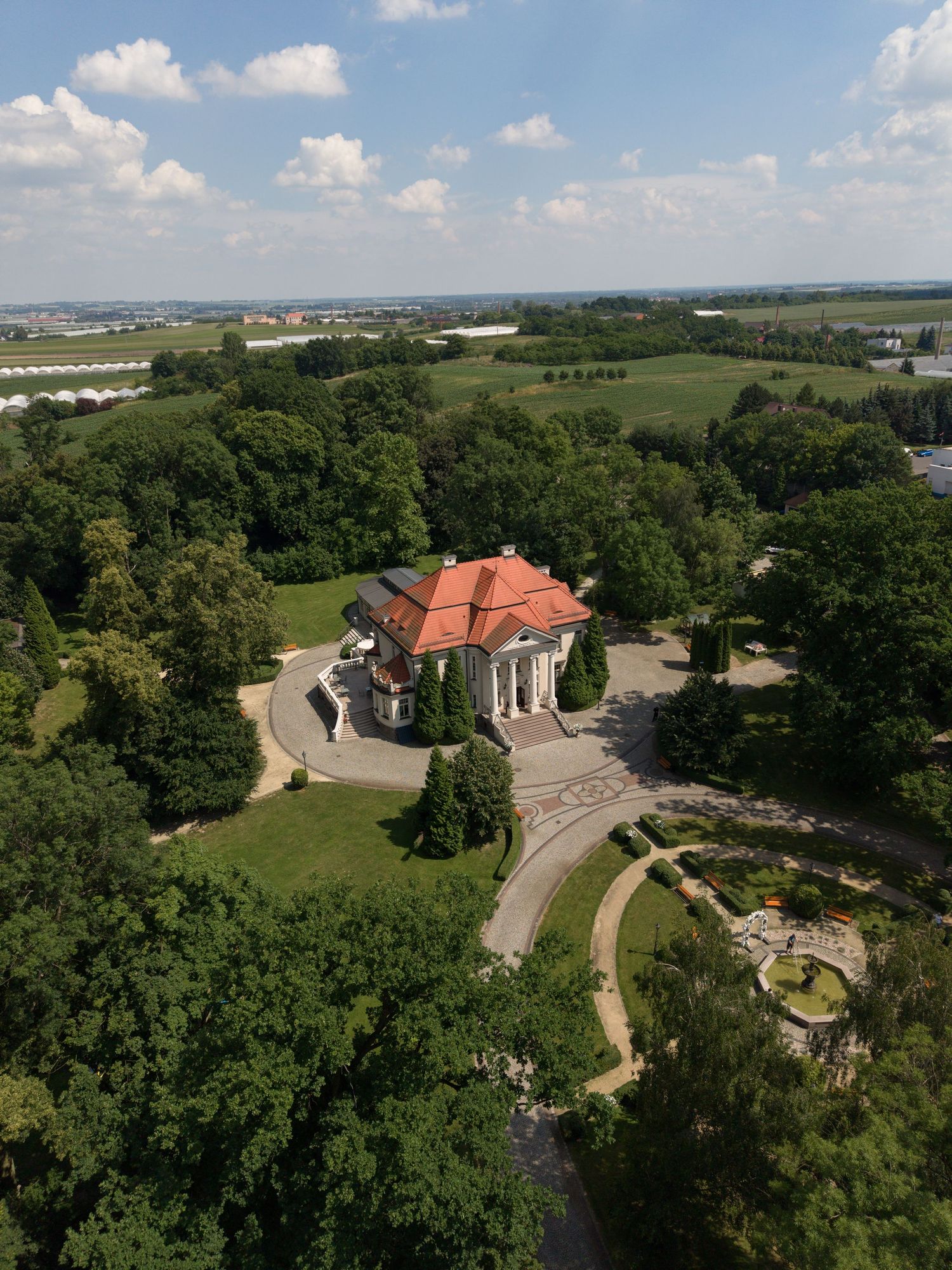 Aerial view of a historic mansion with red roof surrounded by lush green trees and manicured gardens on a sunny day.