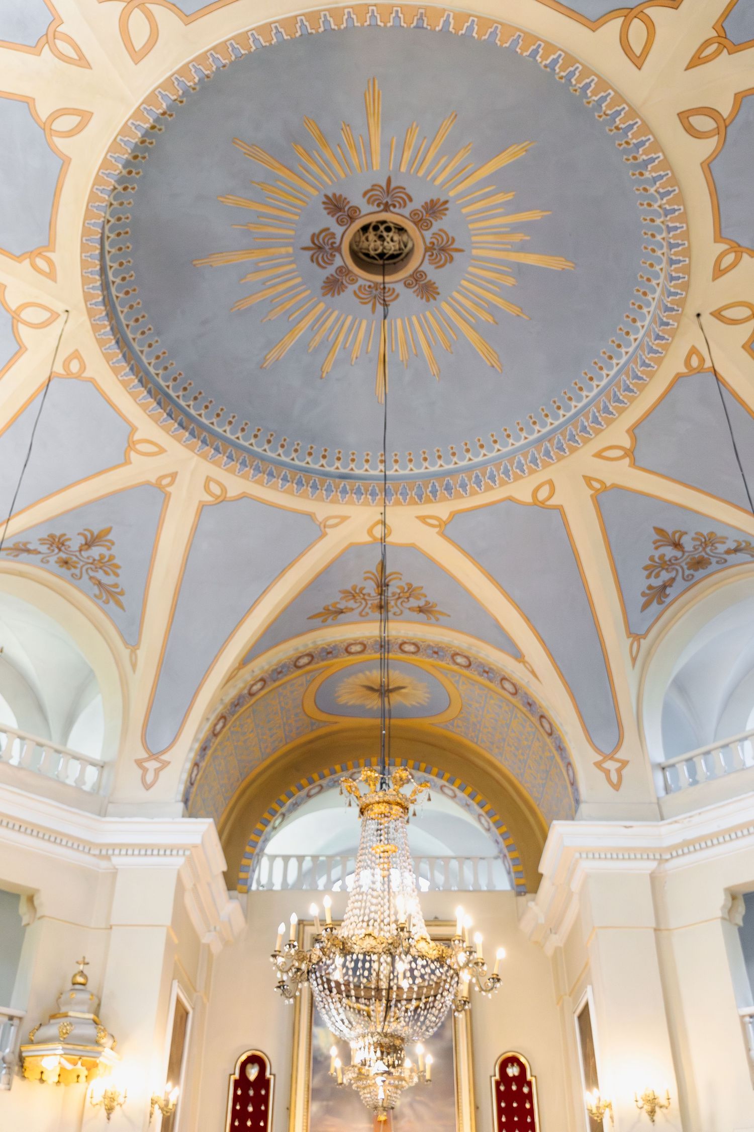 Ornate cathedral ceiling with a golden sunburst medallion, crystal chandelier, and arched white walls with architectural details.