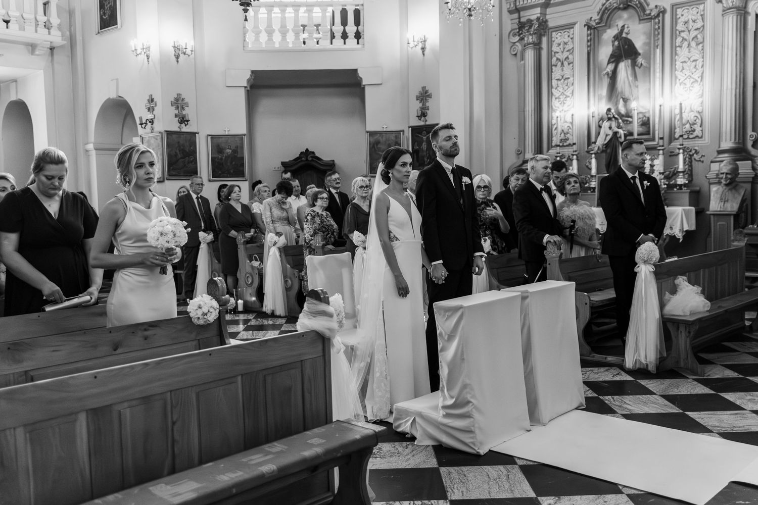 A black and white photograph of a wedding ceremony inside an ornate church with guests seated in wooden pews.