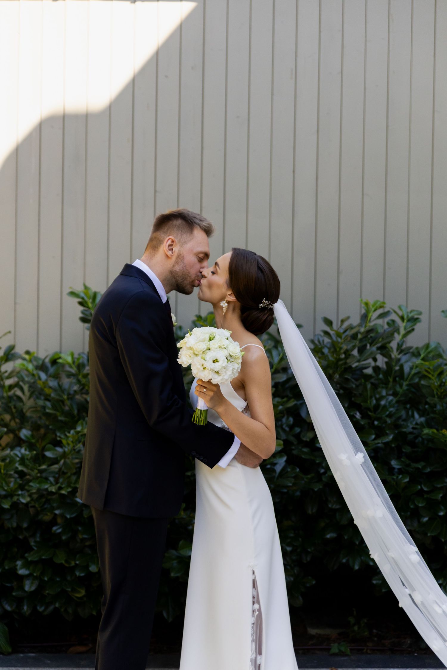 A romantic wedding moment showing a couple kissing while holding a white bouquet, with a flowing veil and greenery backdrop.