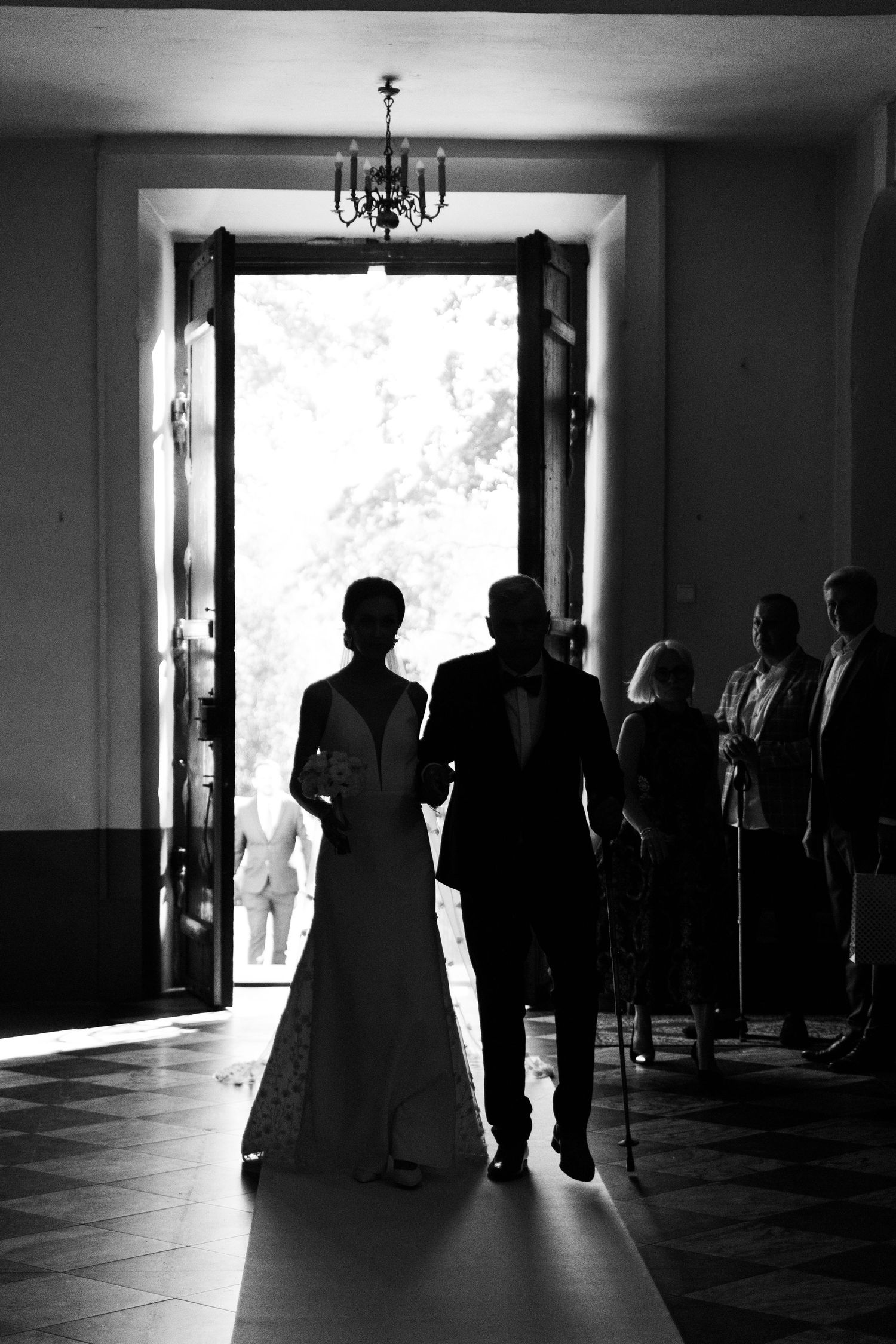 A black and white silhouette of a couple in wedding attire standing in a doorway with chandelier above.