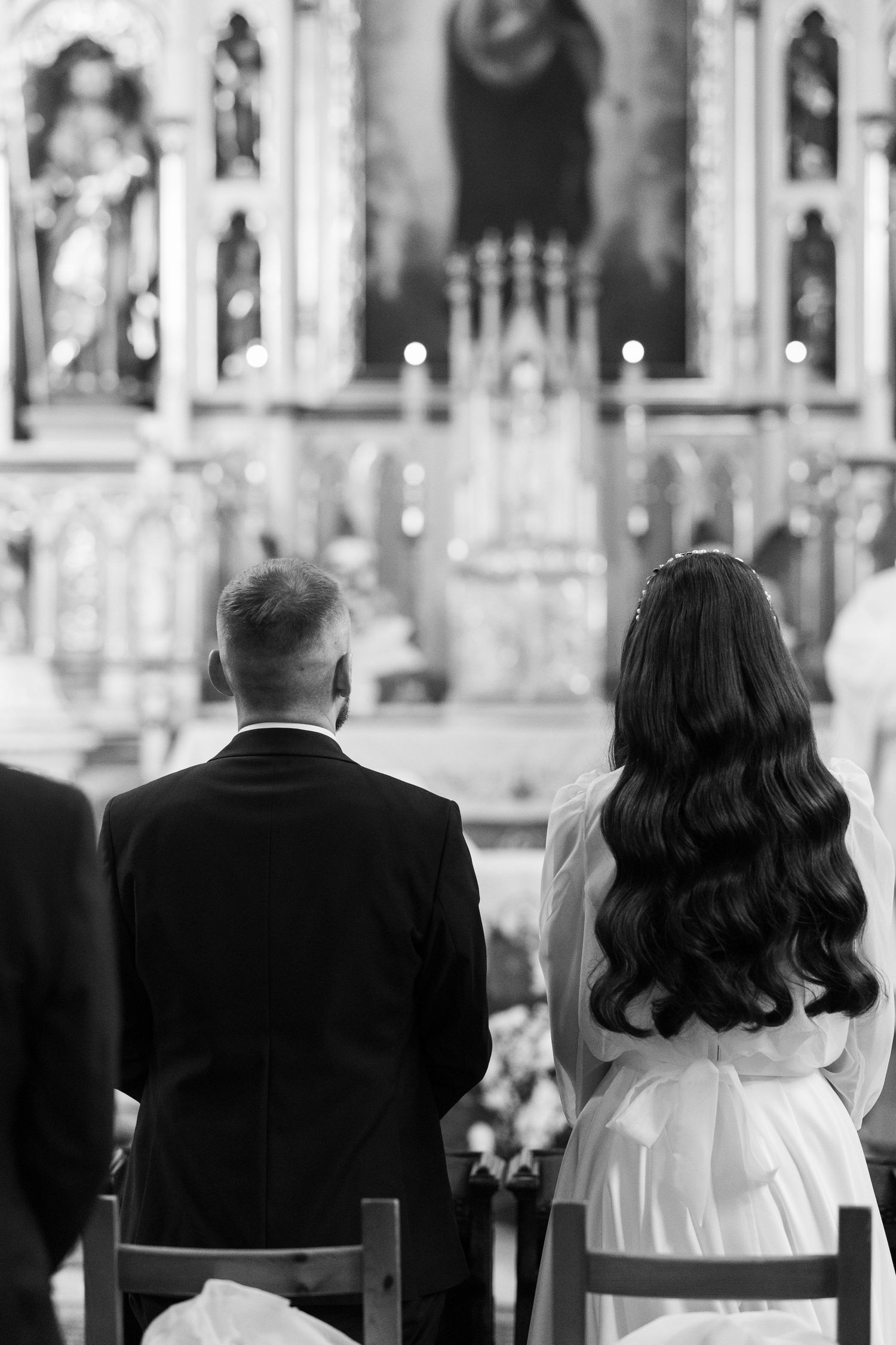 A couple sits in a church during a wedding ceremony, with the bride's dark wavy hair cascading down her back.