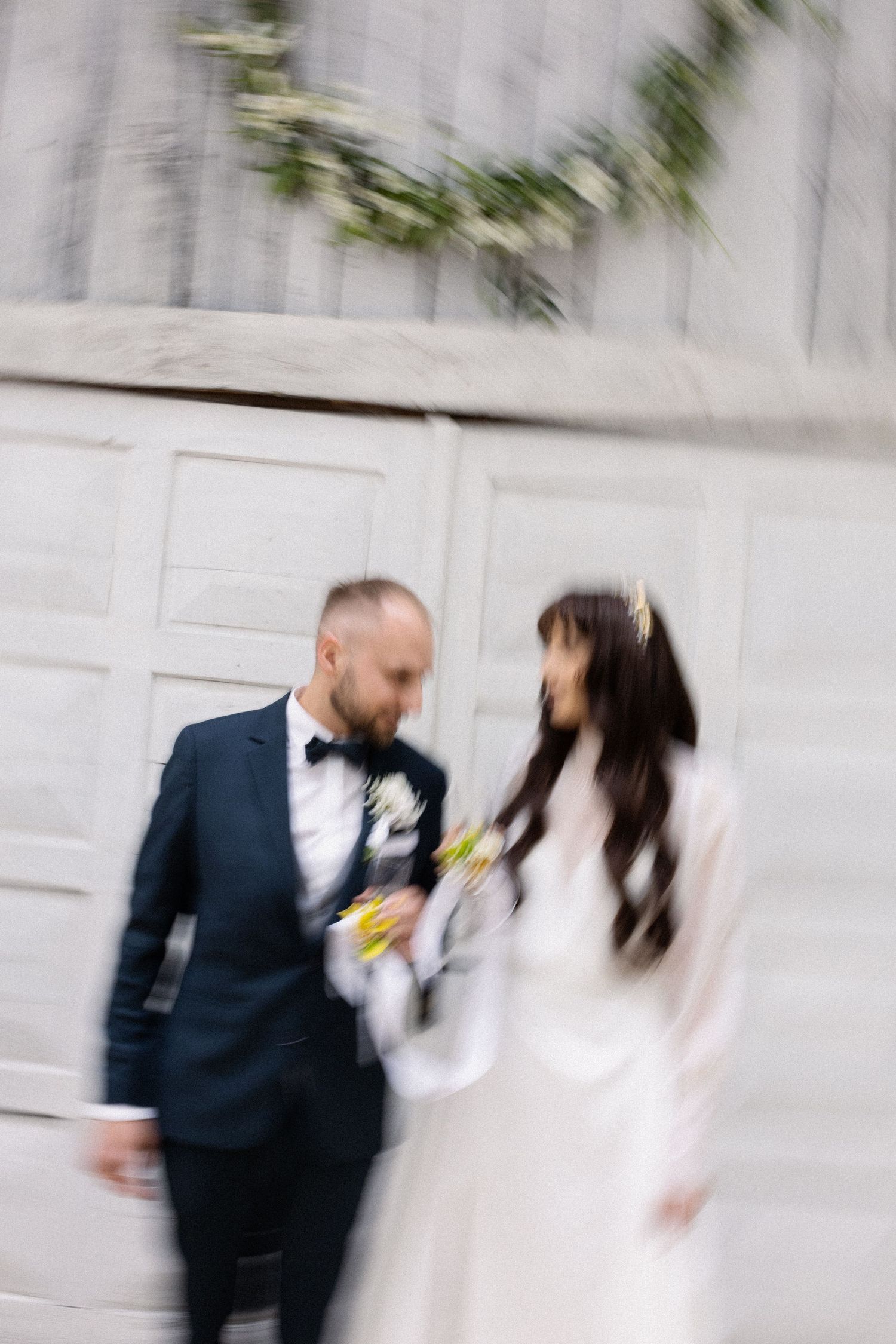 Motion blurred photo of newlyweds in formal attire walking together against a light gray wall with greenery above.