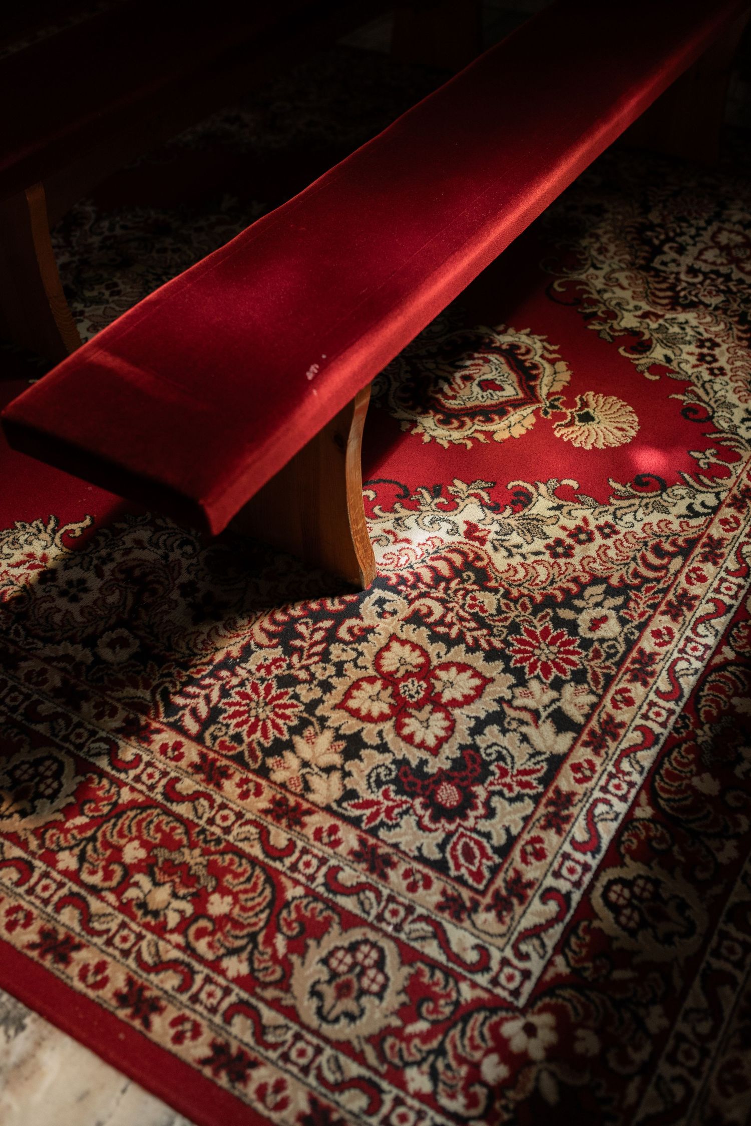 Red velvet prayer bench rests on ornate floral patterned carpet with traditional Persian design in warm lighting.