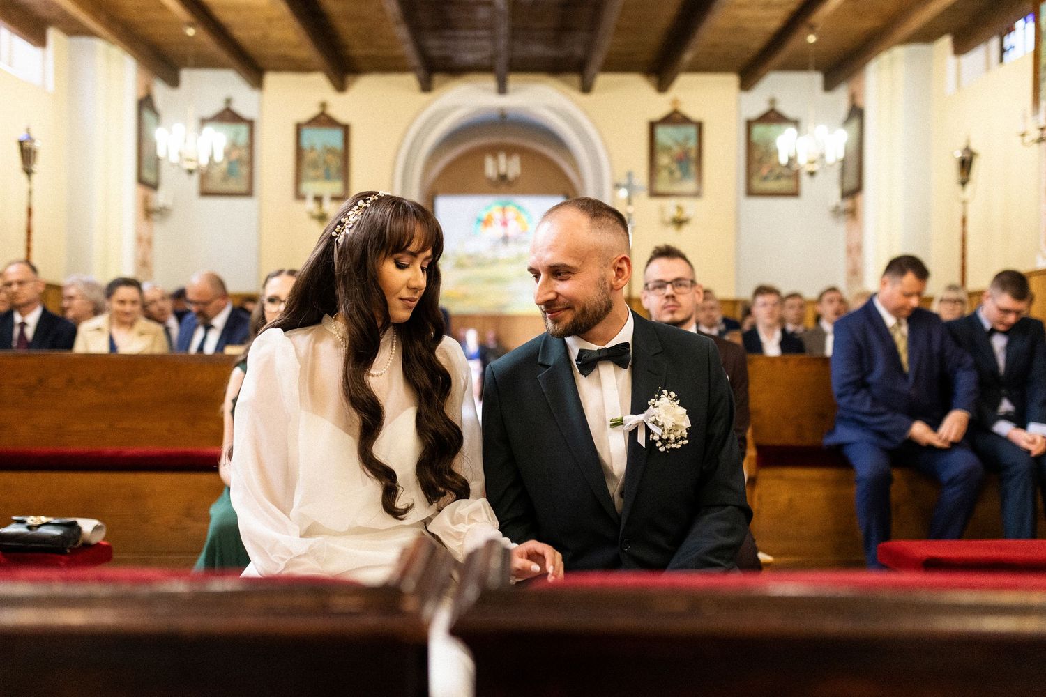 A couple sits together in a church pew during a wedding ceremony with guests visible in the background.
