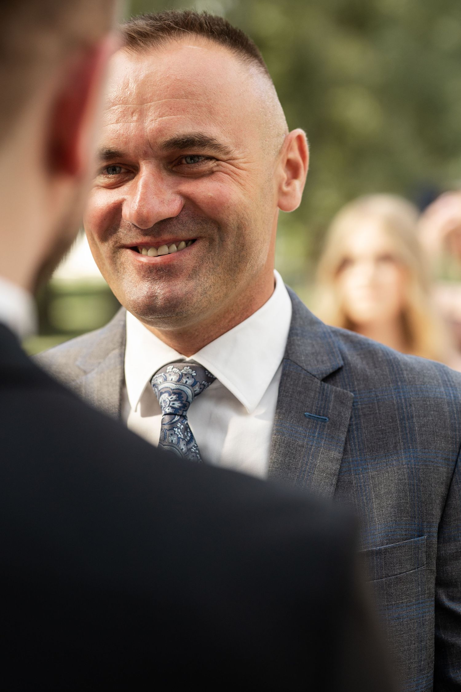 A well-dressed man in a gray plaid suit and patterned tie smiles warmly during a formal outdoor event.