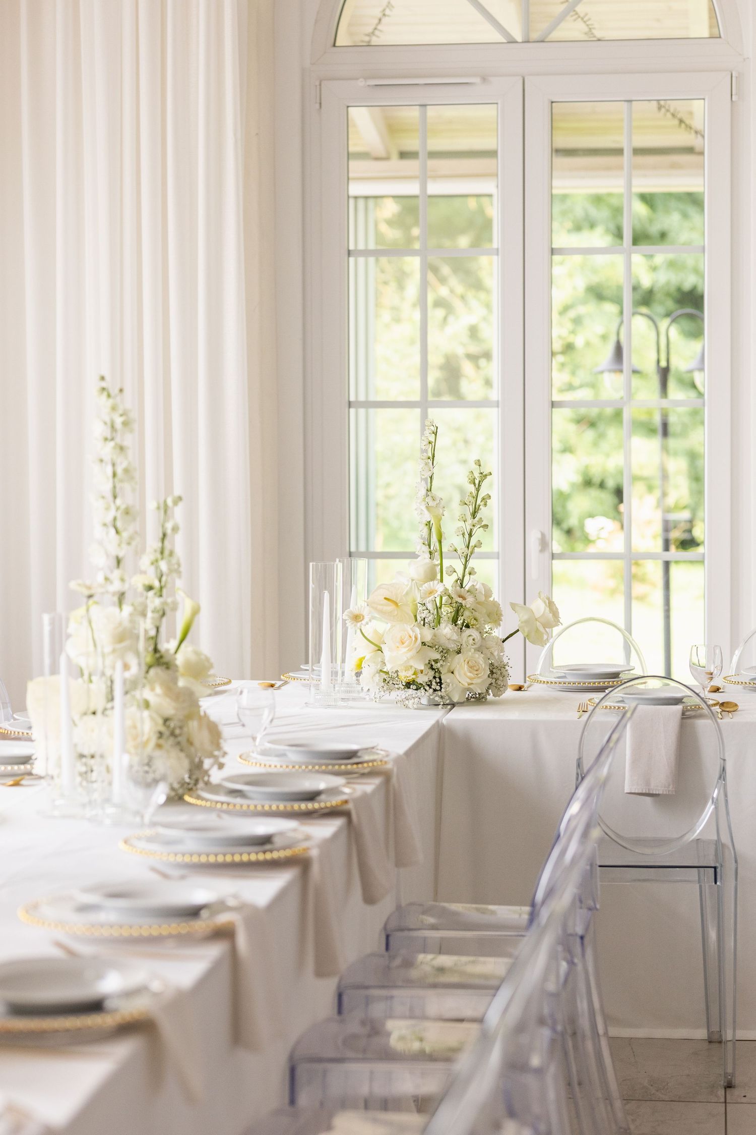 Elegant white dining room with multiple place settings and floral centerpieces illuminated by natural window light.