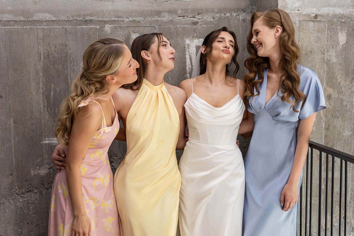 Five friends in pastel dresses pose together against a concrete wall in a candid photo series.