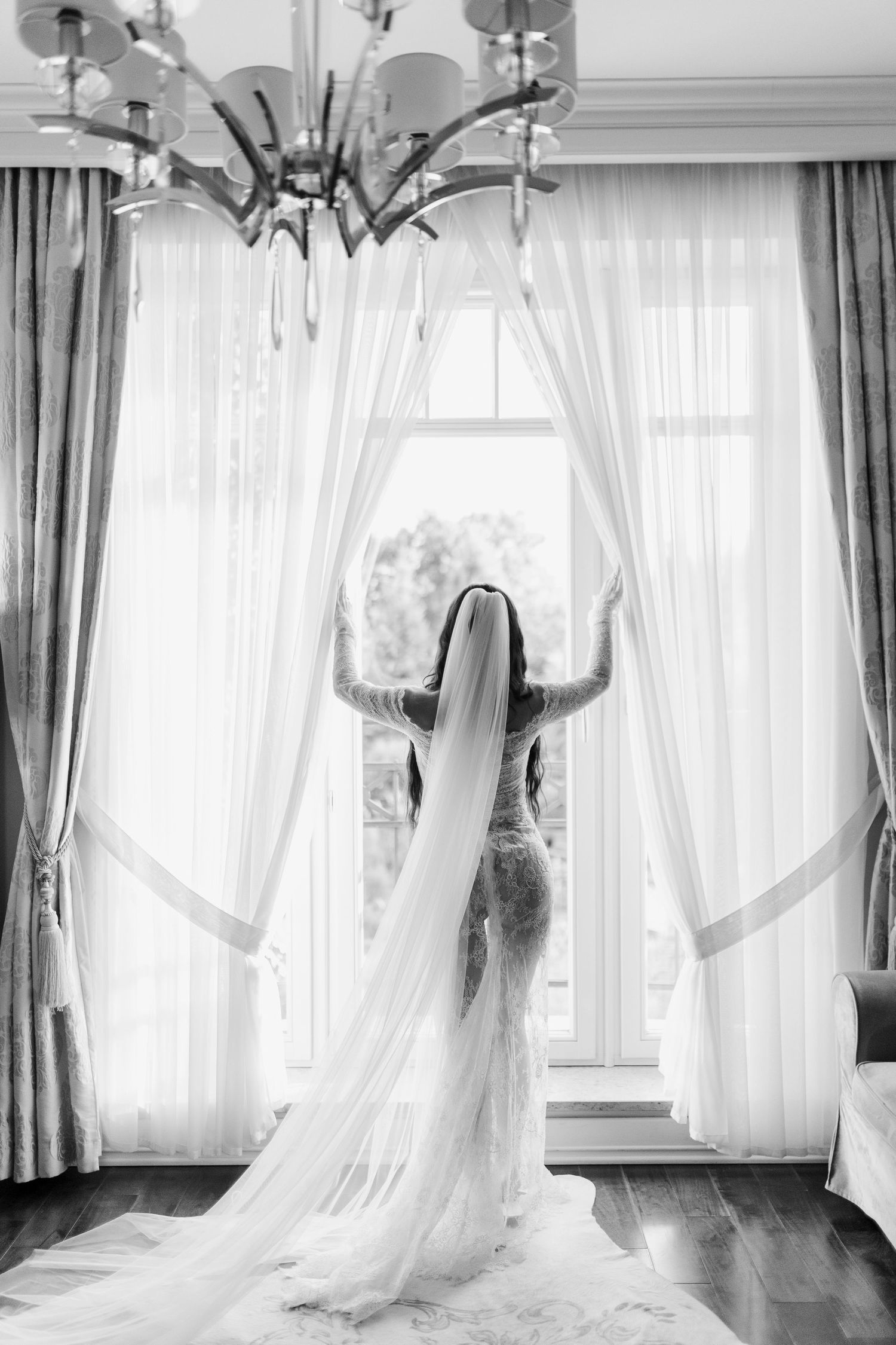 Silhouette of bride in flowing gown and veil opening elegant curtains by window.