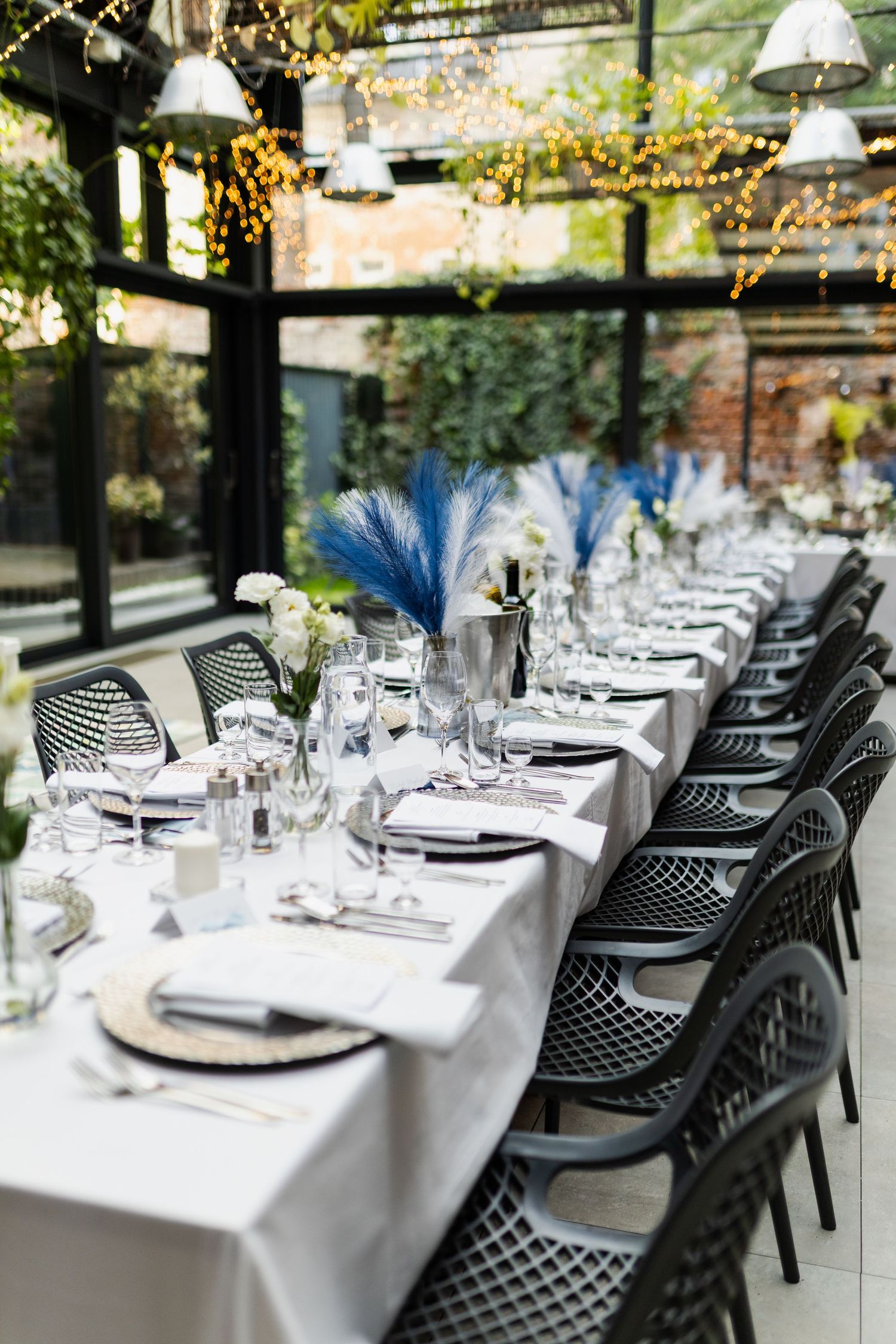 Elegant table setting with blue feather centerpieces, white linens, and modern black chairs in a glass-enclosed dining space.