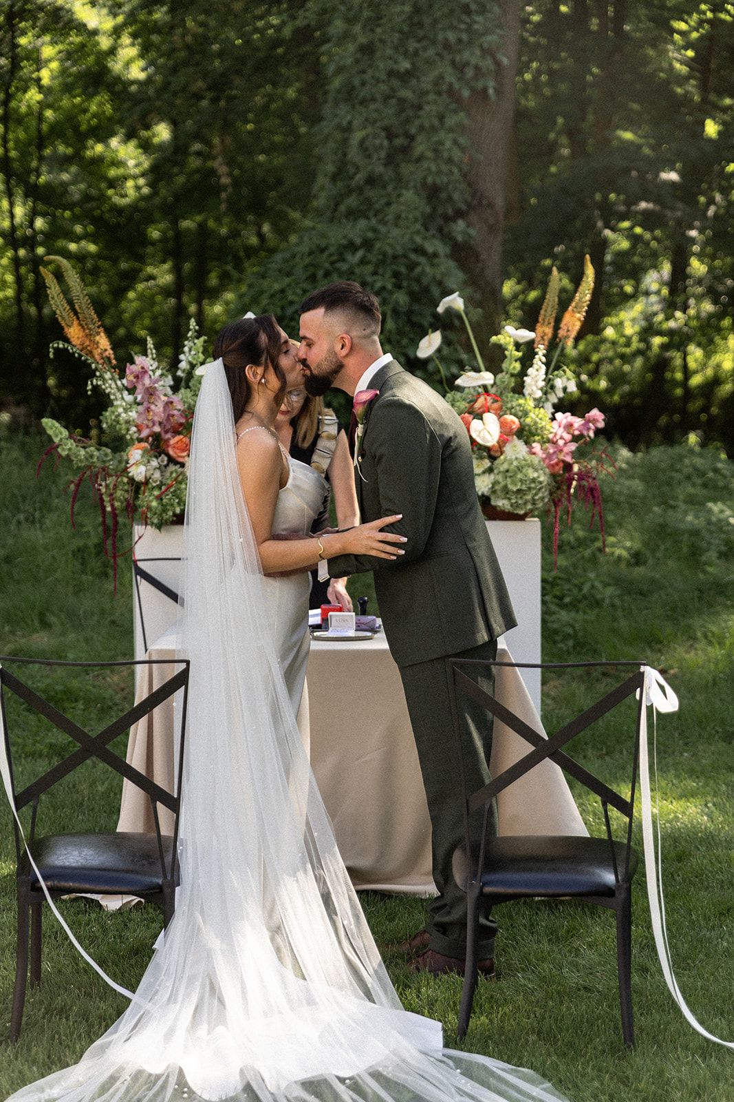A romantic outdoor wedding ceremony shows a couple sharing their first kiss as newlyweds in a garden setting.