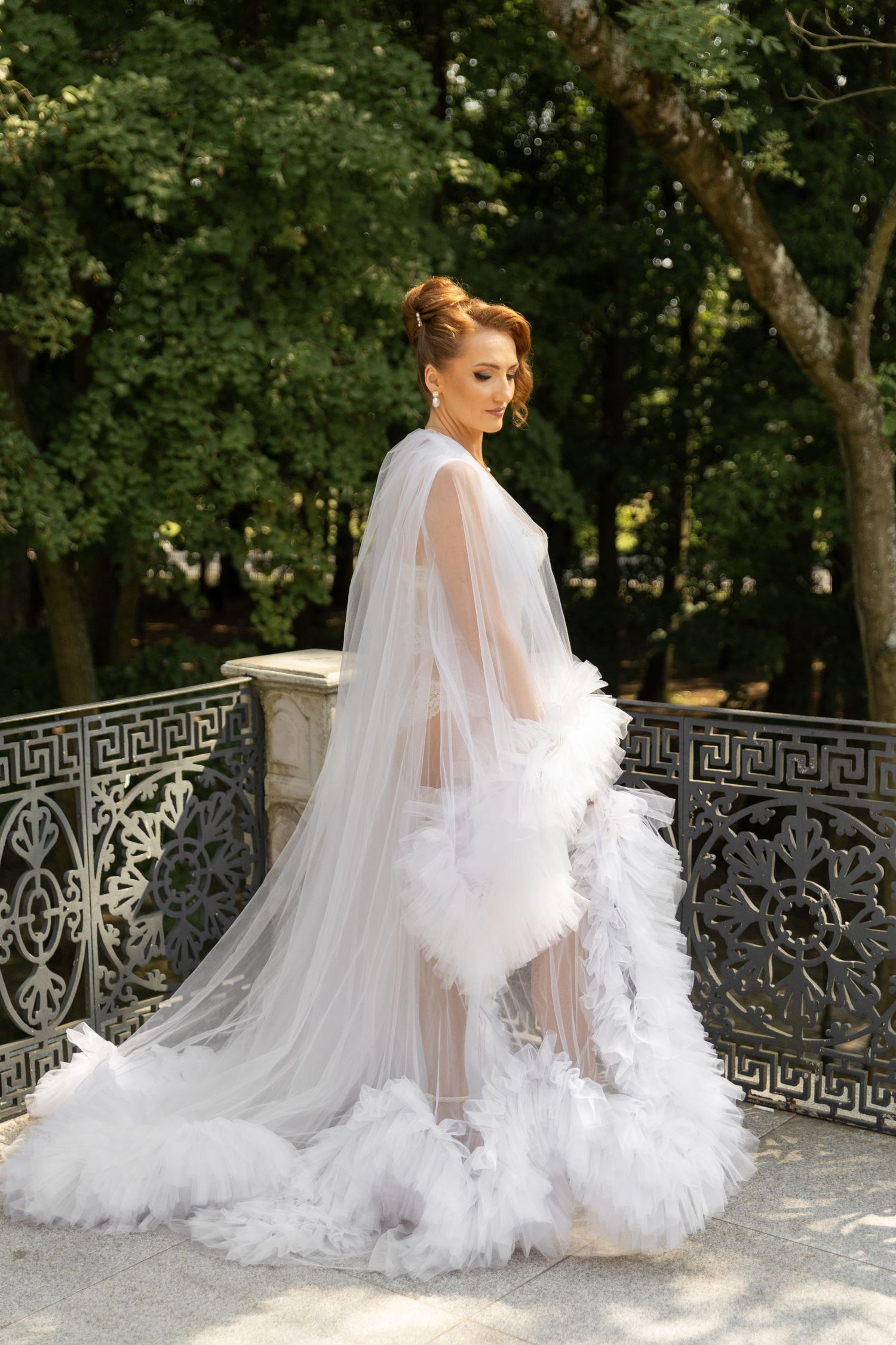 A flowing white bridal boudoir robe with feather trim is showcased in an elegant outdoor garden setting.